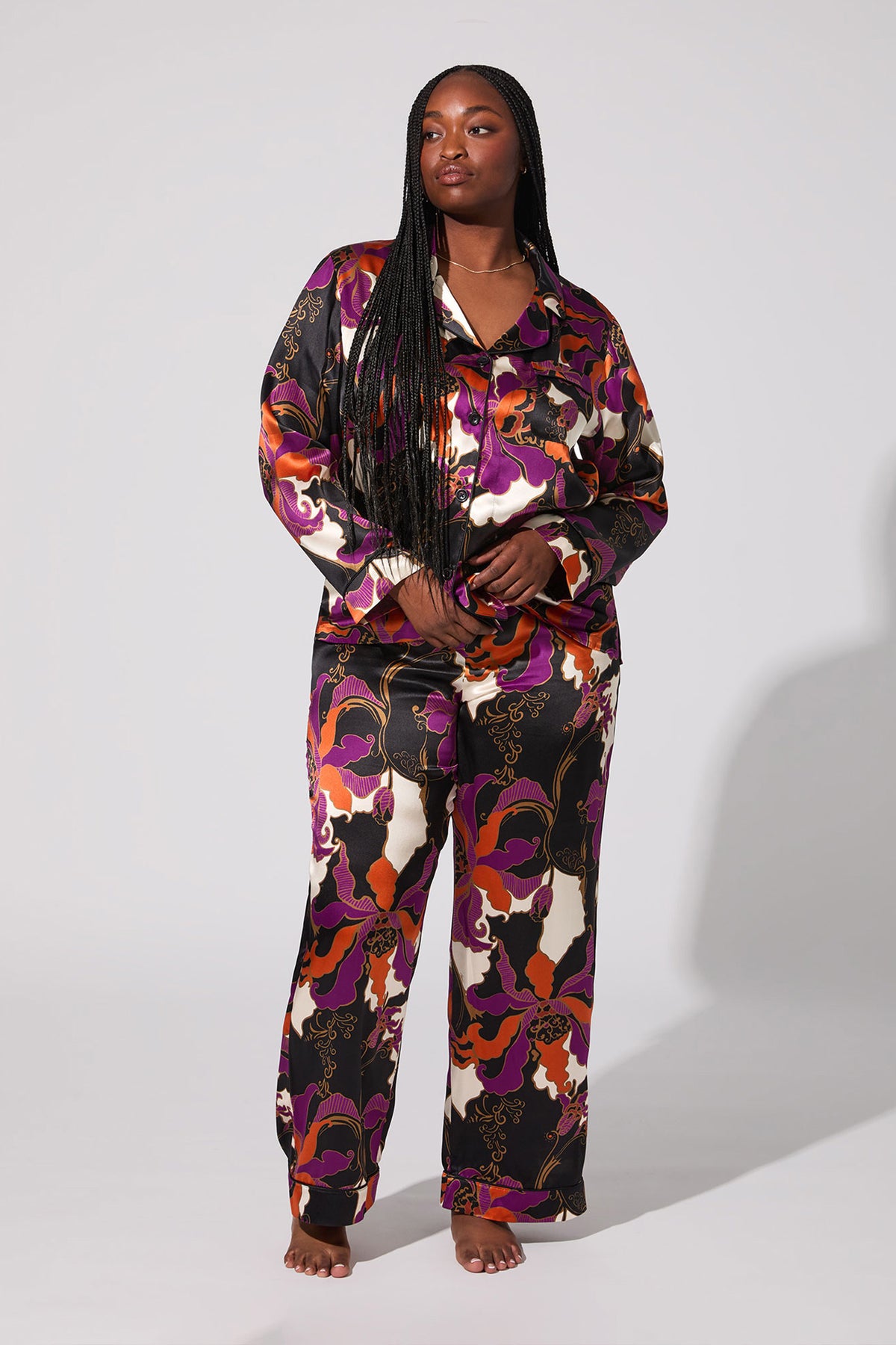 A woman with long braided hair stands barefoot and confidently in the Fall Floral Classic Silk Pj, featuring a bold purple, orange, red, black, and cream floral pattern, against a plain background.