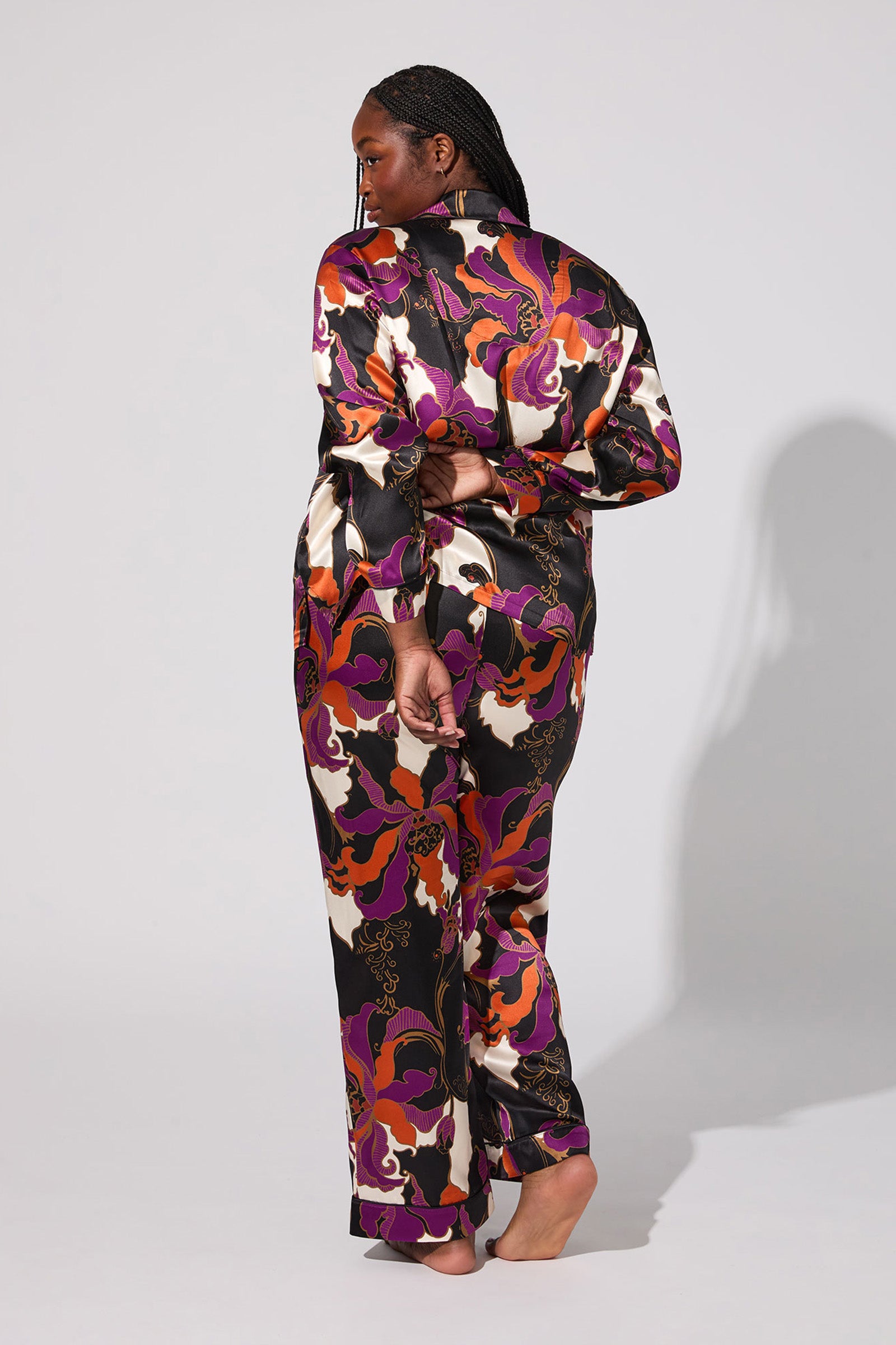 A person with braided hair stands barefoot, facing away, wearing the Fall Floral Classic Silk Pj—a long-sleeve satin pajama set with a vibrant abstract floral pattern in purple, orange, black, and white. Shadow falls to their right on a plain background.