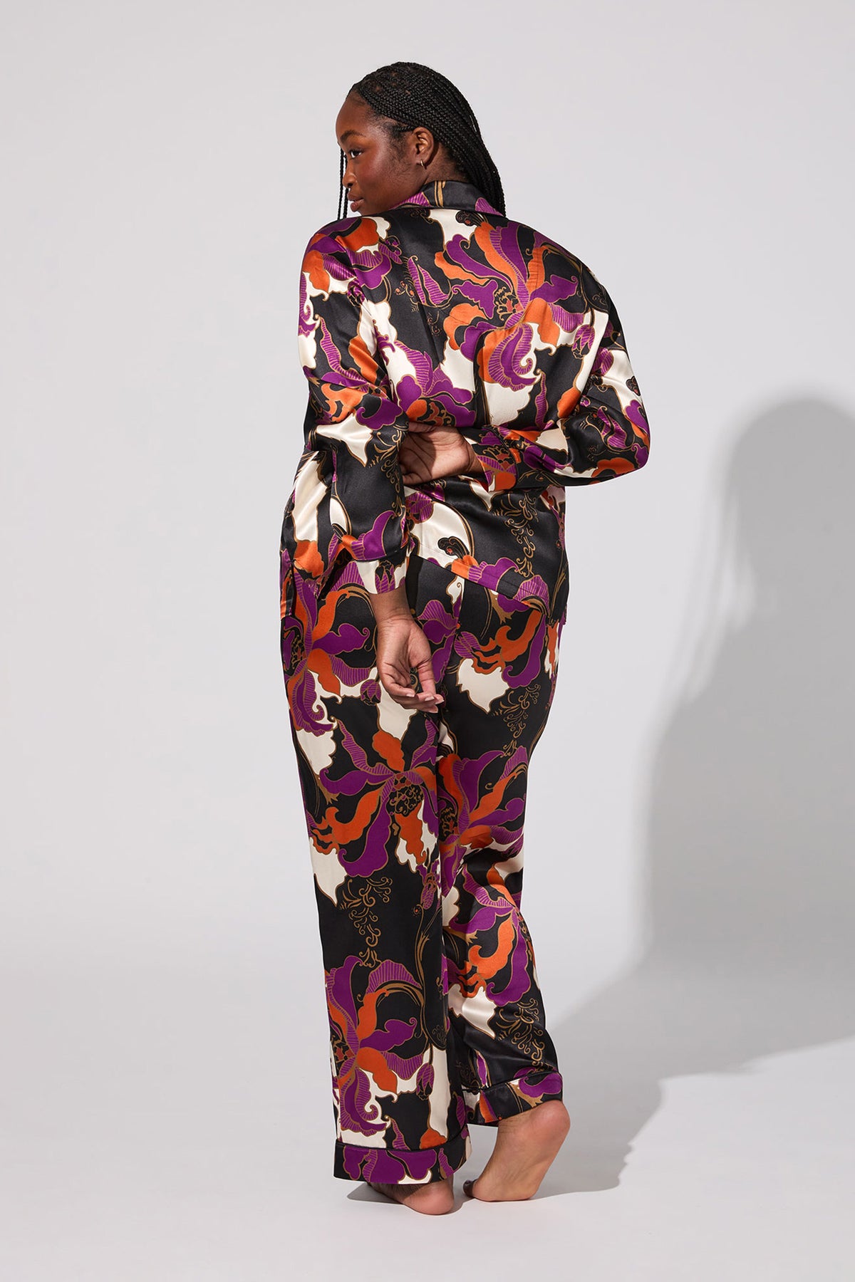 A person with braided hair stands barefoot, facing away, wearing the Fall Floral Classic Silk Pj—a long-sleeve satin pajama set with a vibrant abstract floral pattern in purple, orange, black, and white. Shadow falls to their right on a plain background.