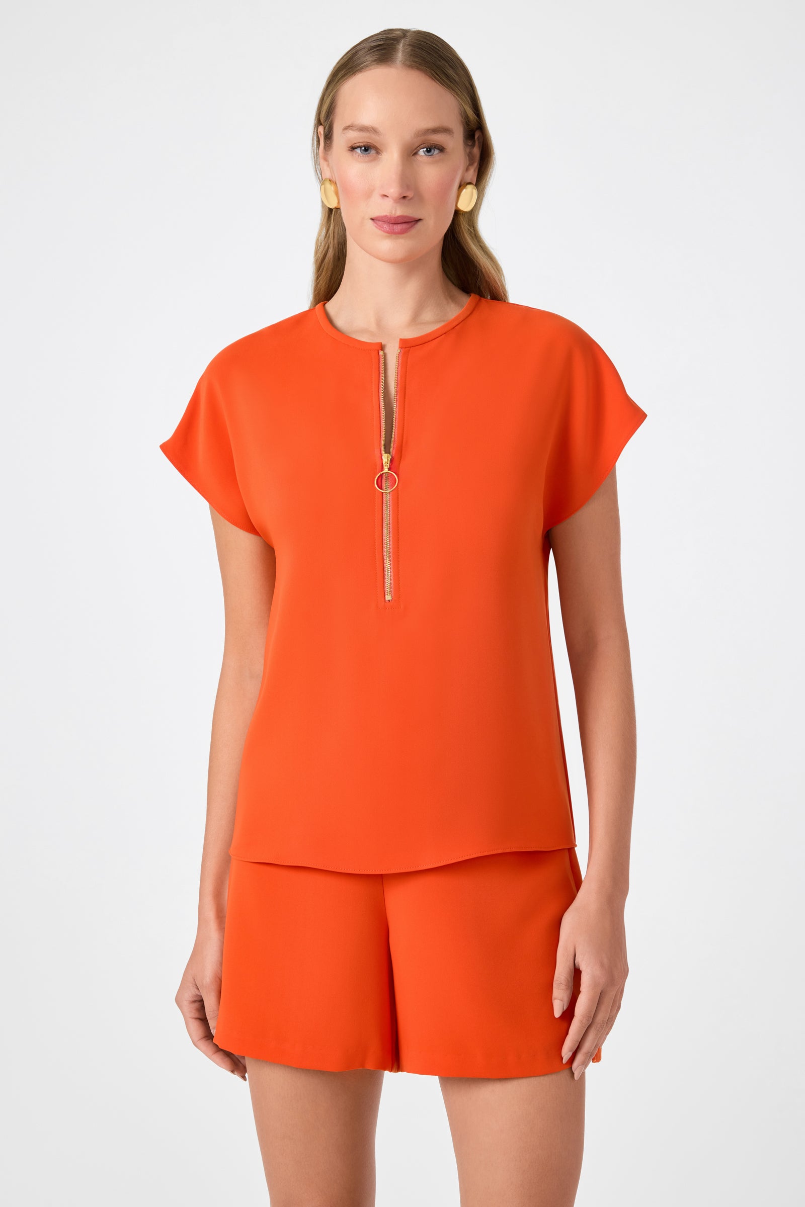 A woman with long blonde hair and gold hoop earrings wears the Hasil Top - Double Georgette, a bright orange short-sleeve top with a front zipper, paired with matching shorts against a plain light background.