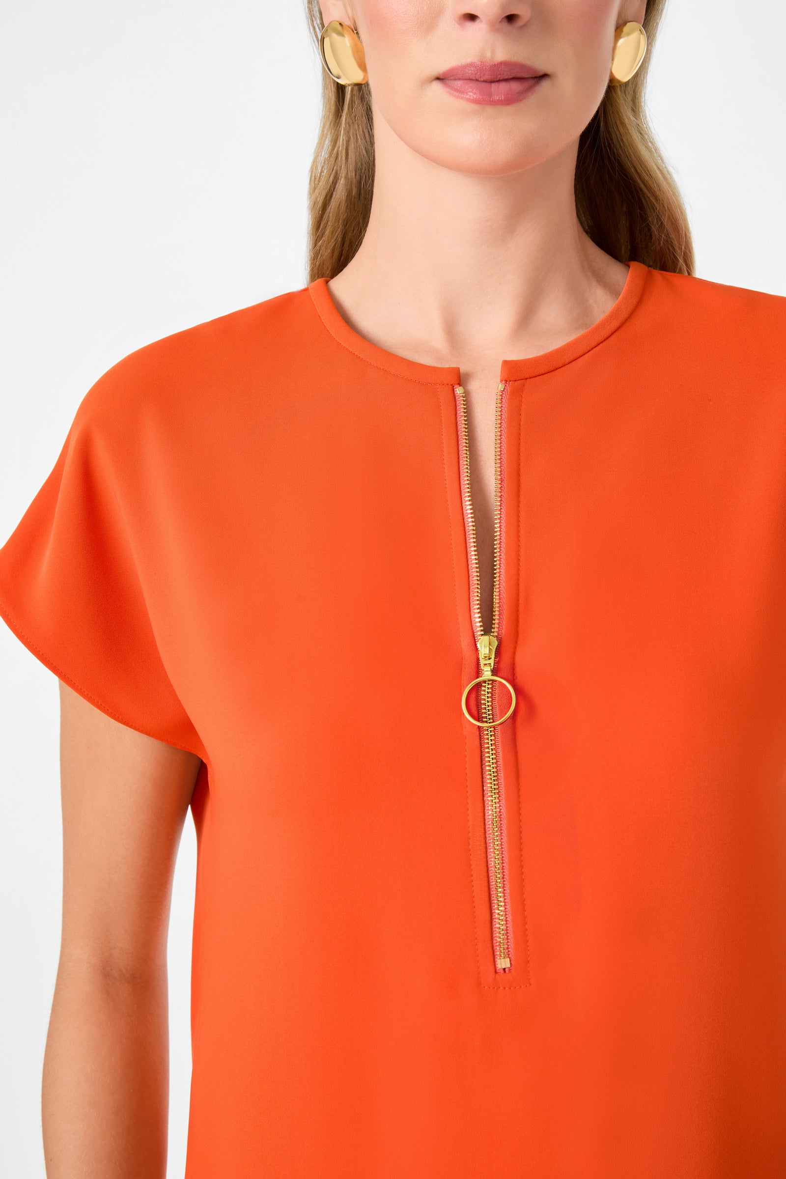 A woman wears the Hasil Top - Double Georgette in bright orange, featuring short sleeves and a front gold zipper with a round pull ring. She accessorizes with gold hoop earrings; the image shows her from shoulders to above the waist.