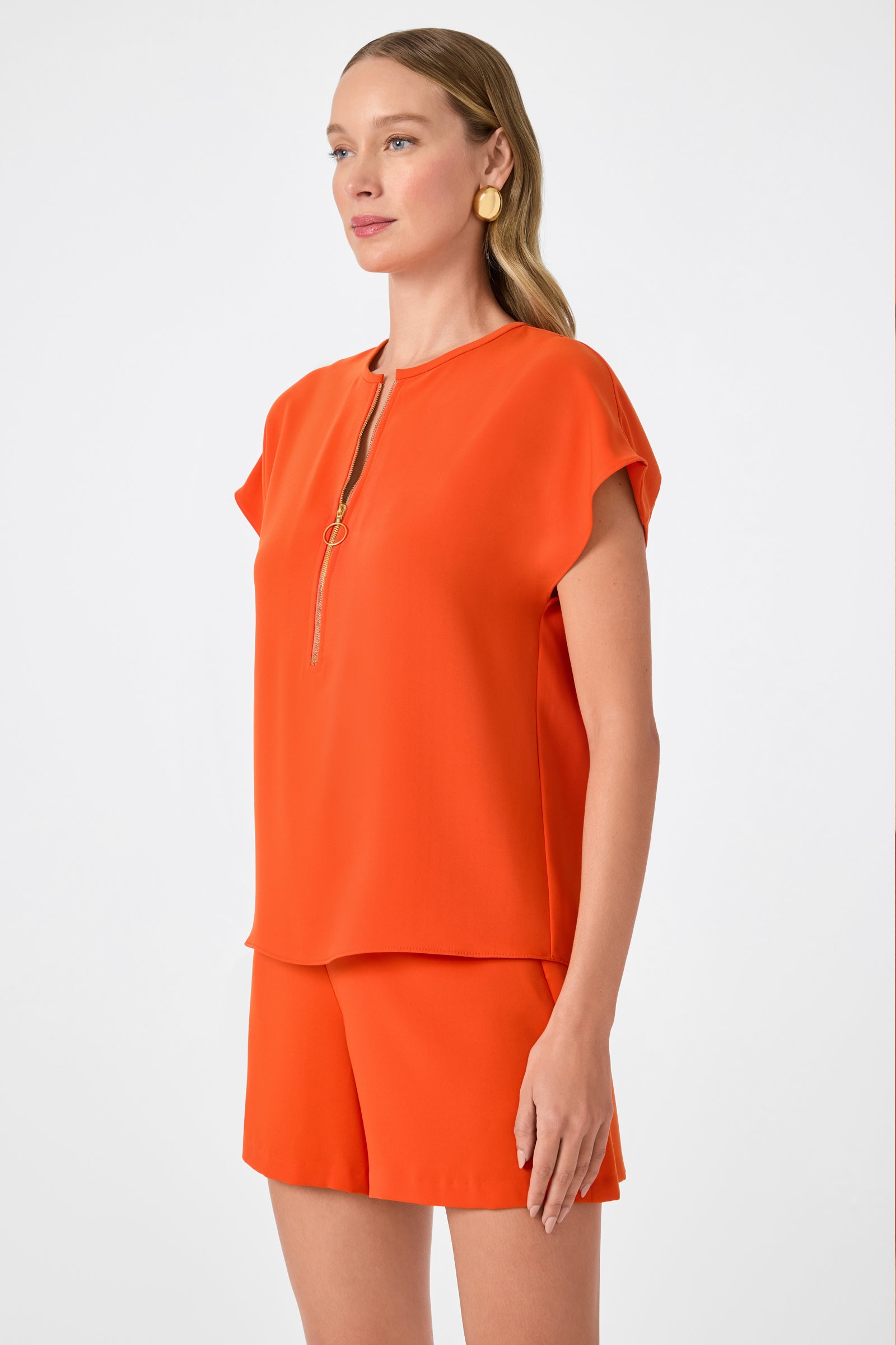 A woman stands against a white background wearing the Hasil Top - Double Georgette in bright orange with short sleeves and a front zipper, paired with matching shorts. She has straight light brown hair and gold circular earrings.
