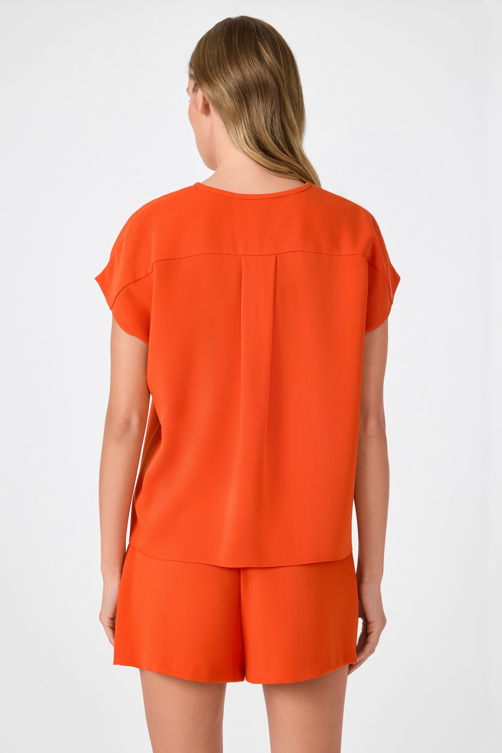 A woman with long, light brown hair stands facing away, wearing the Hasil Top - Double Georgette in a loose-fitting orange style with matching shorts against a plain light background.