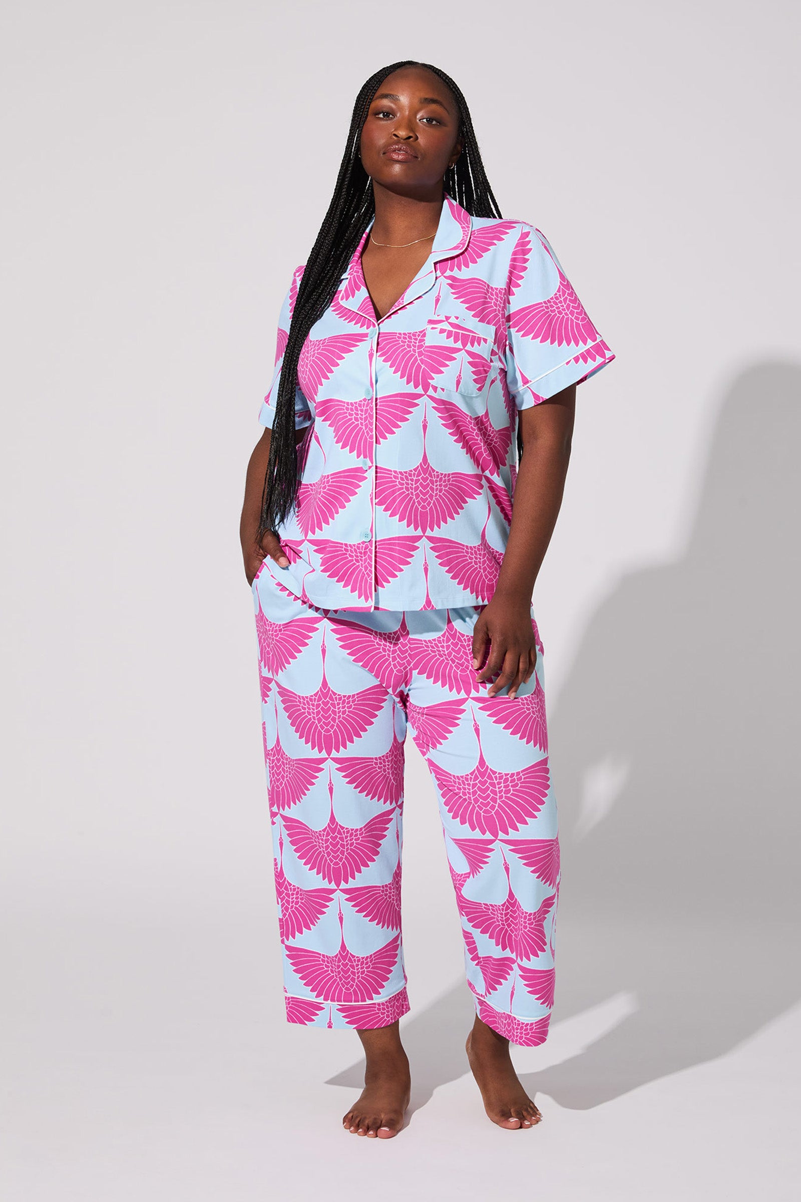 A woman stands barefoot against a plain background, wearing the Cranes Crop Pj Set—a light blue pajama set with a bold pink bird pattern, featuring a short-sleeve button-up shirt and matching pants.