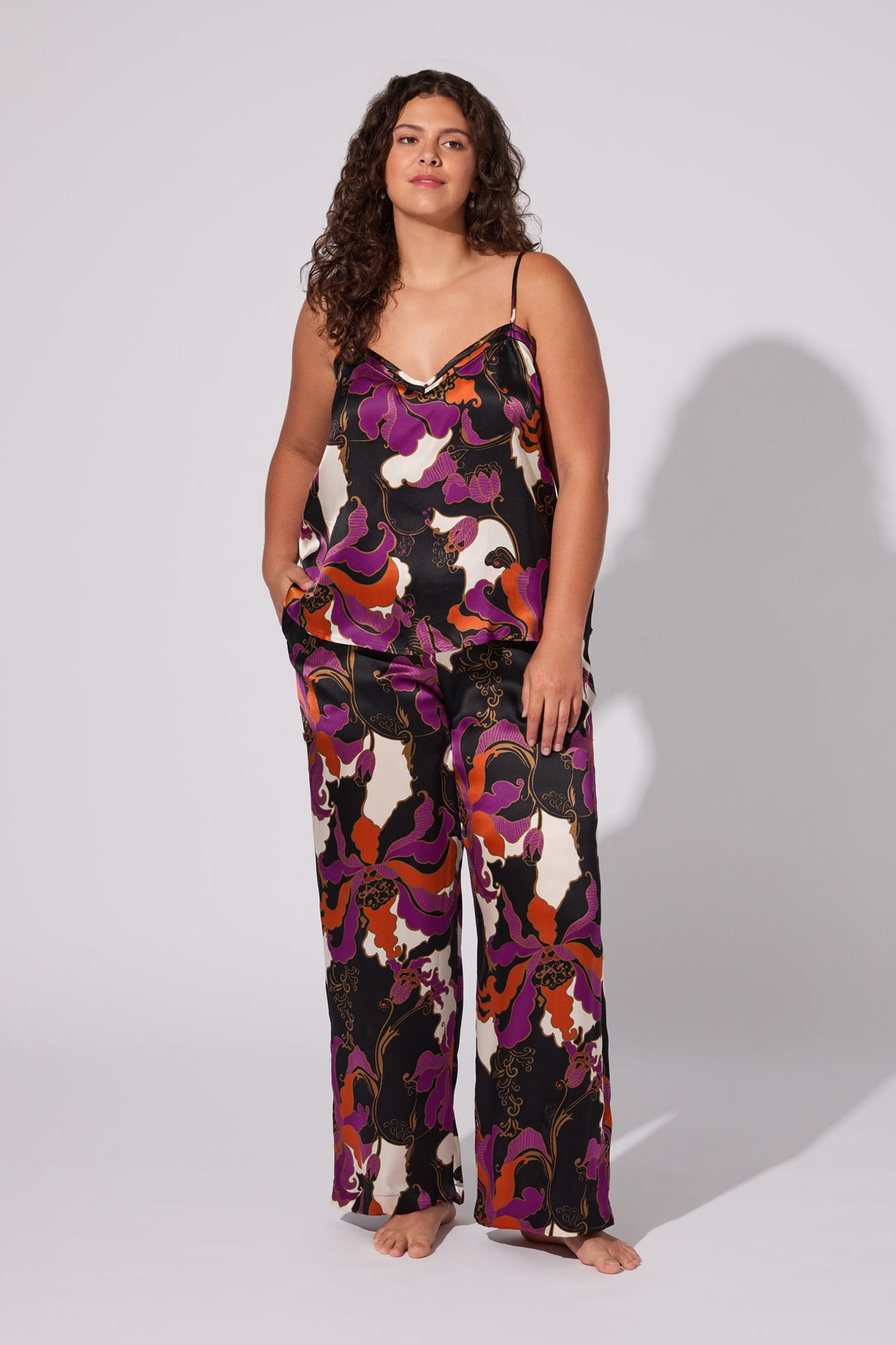 A woman with long curly hair models the Fall Floral Silk Cami Pj Set—colorful, loose-fitting camisole and matching wide-leg pants with a bold floral pattern—standing barefoot against a plain, light background.
