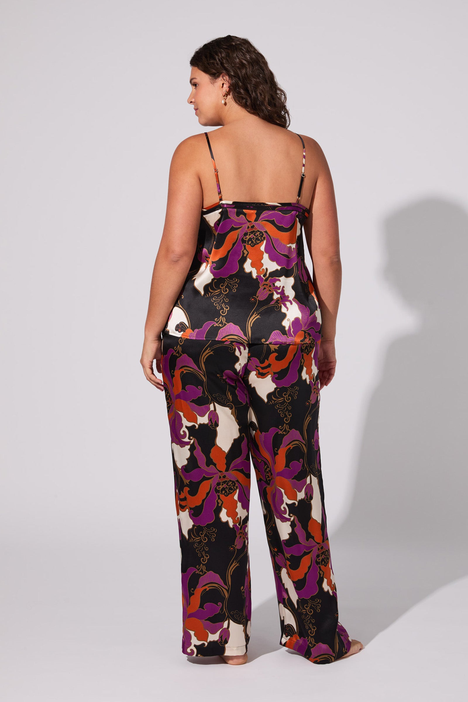 A woman with wavy brown hair stands barefoot, facing away, wearing the Fall Floral Silk Cami Pj Set featuring a colorful floral and abstract pattern on a plain light gray background.