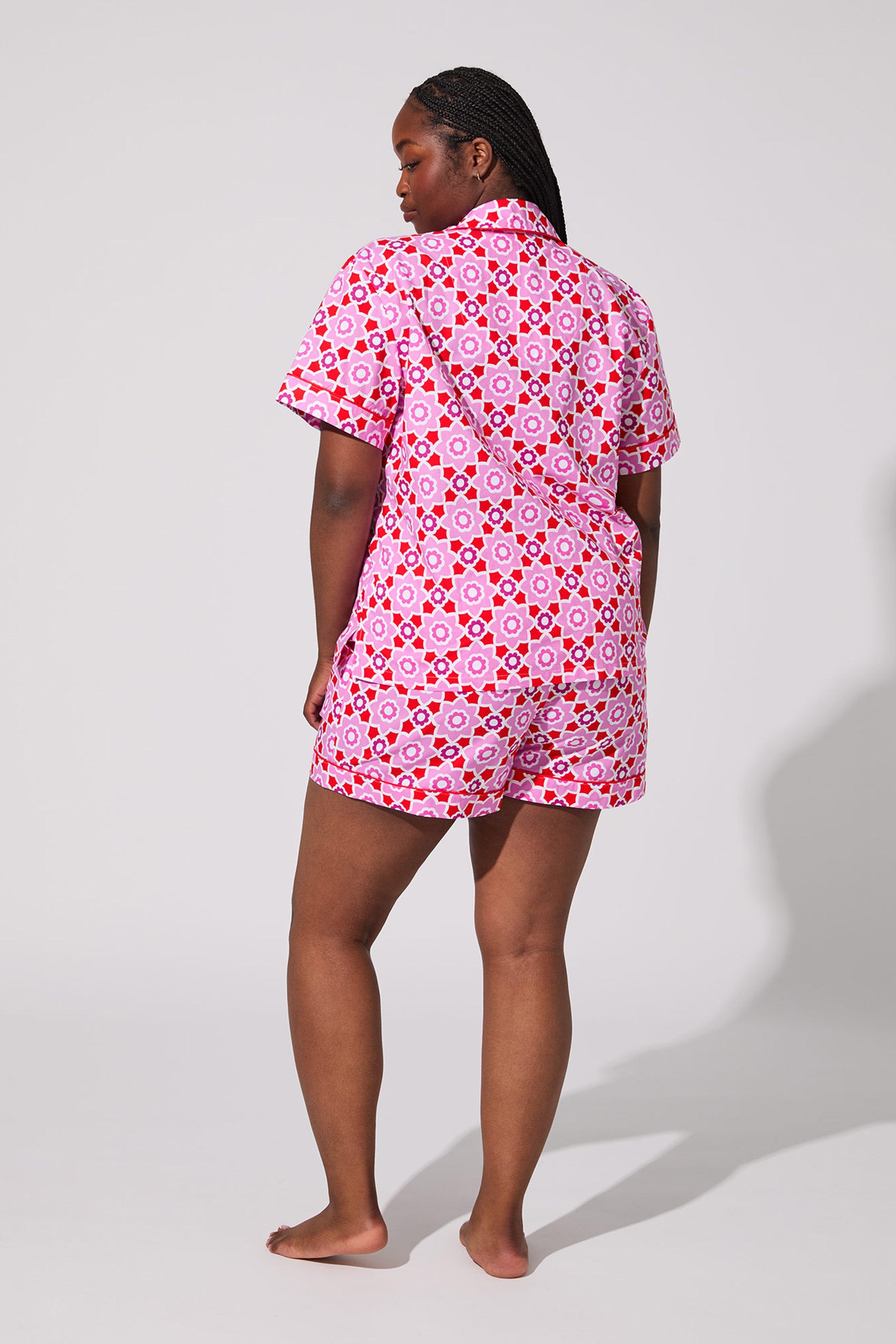A person stands barefoot facing away, wearing the Mosiac Tile Short Pj Set—a pink and red patterned short-sleeve shirt and shorts—against a plain light gray background.