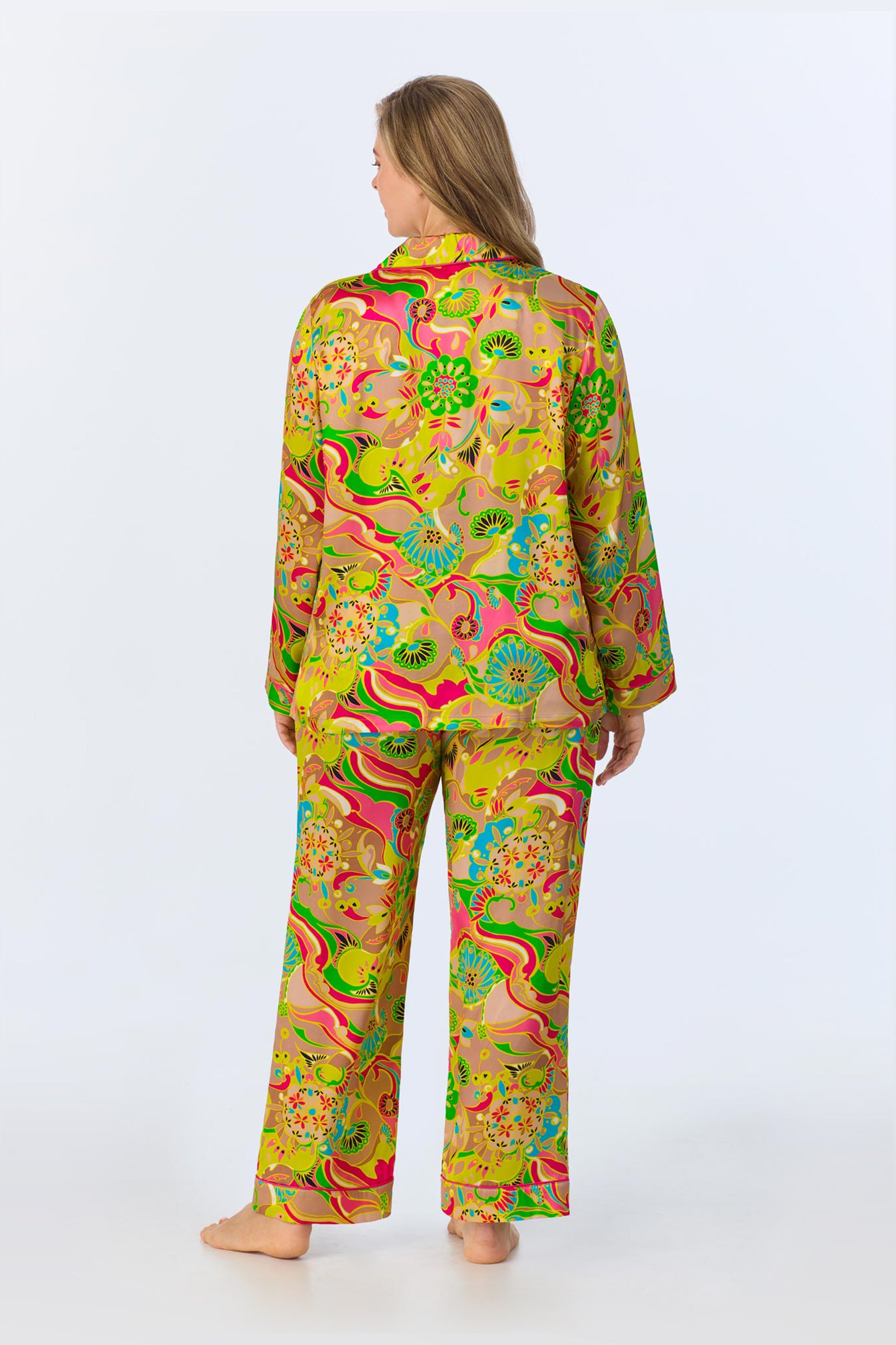 A person stands barefoot facing away, wearing the TT LS Pant PJ Set - Heritage Silk, featuring colorful, loose-fitting pajamas with a vibrant floral and abstract pattern in yellow, green, pink, and blue on a light background.