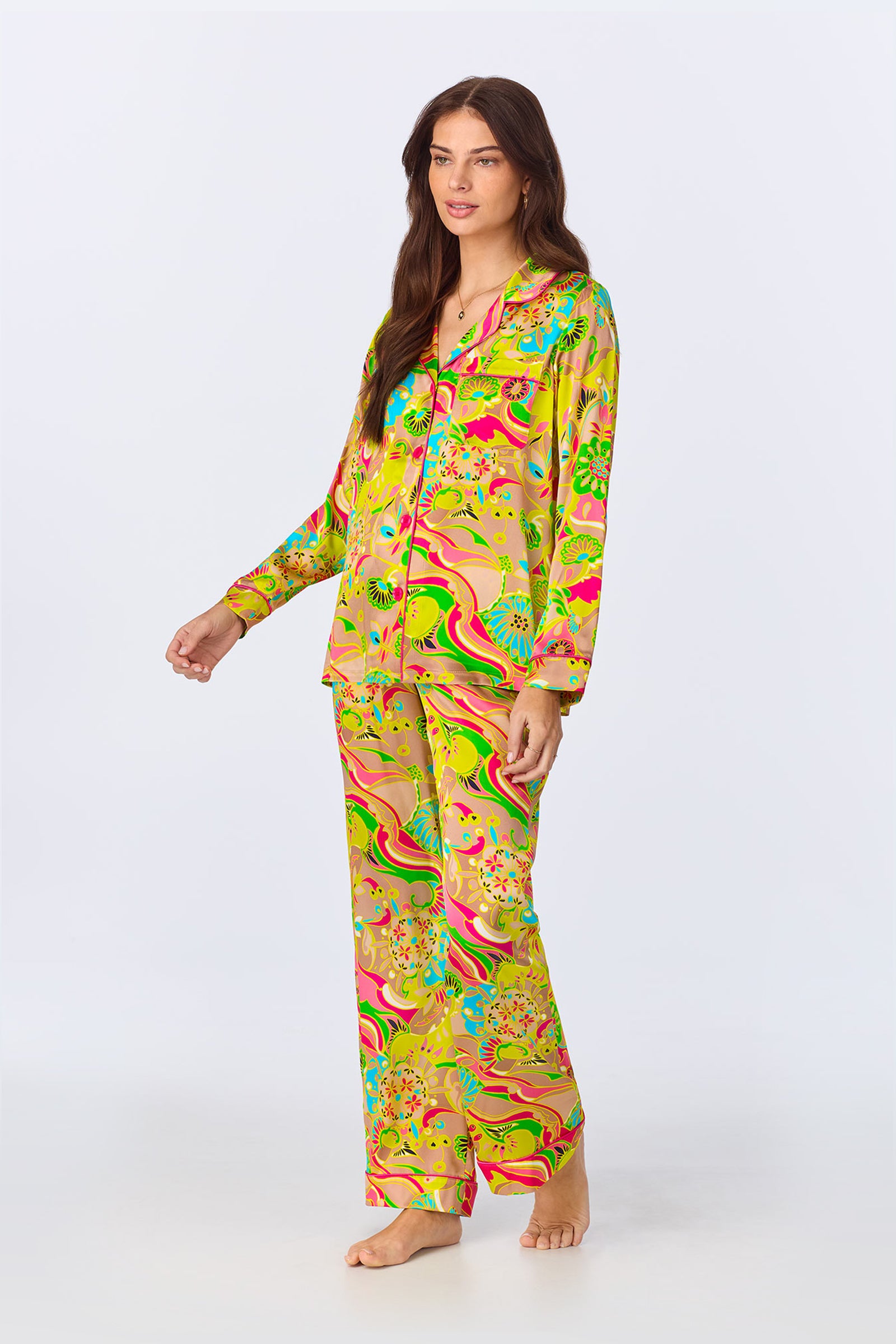 A woman stands barefoot in the TT LS Pant PJ Set - Heritage Silk, featuring bright, colorful satin with a swirling floral and abstract pattern. The set includes a long-sleeved button-up shirt and matching pants against a plain white background.