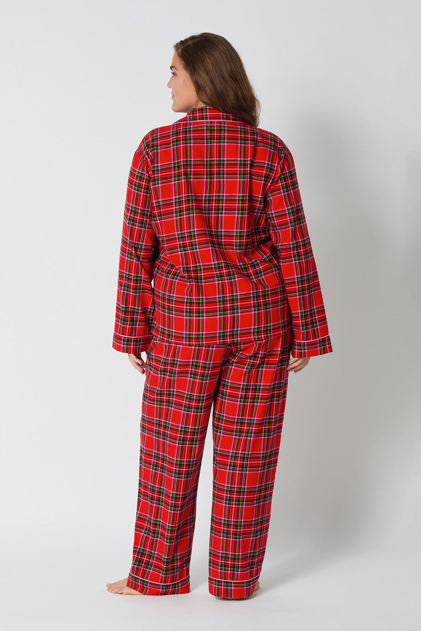 A person stands barefoot facing away against a plain light background, wearing the Christmas Plaid L/S Classic Pj set featuring red, black, green, and white plaid with long sleeves and long pants.