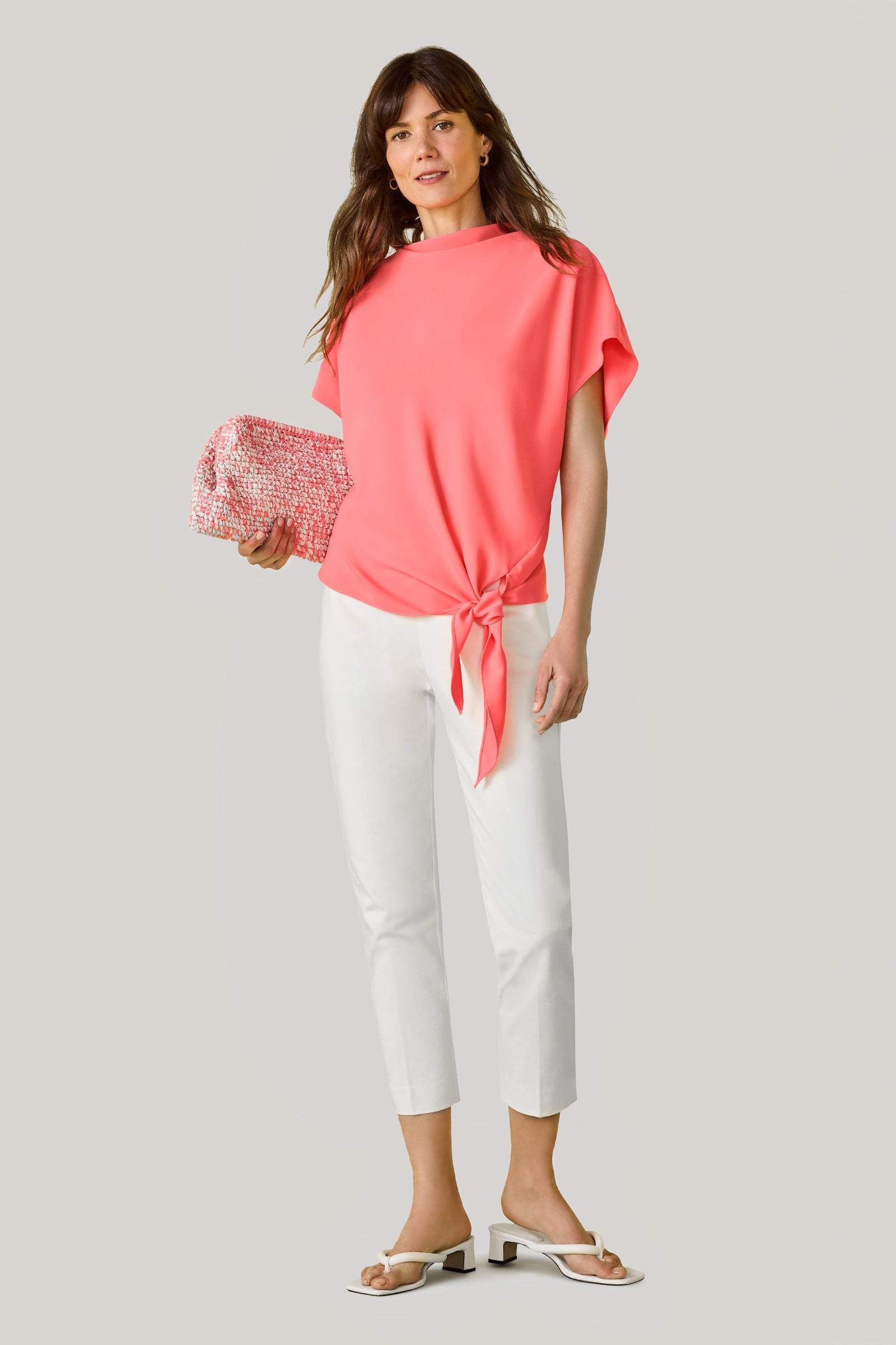 A woman wears a loose coral short-sleeve top with a front tie, the Tie Breaker Slim Crop Pant - Solid Suiting in white, and white sandals. She holds a pink and white clutch, has wavy brown hair, and stands against a plain light gray background.