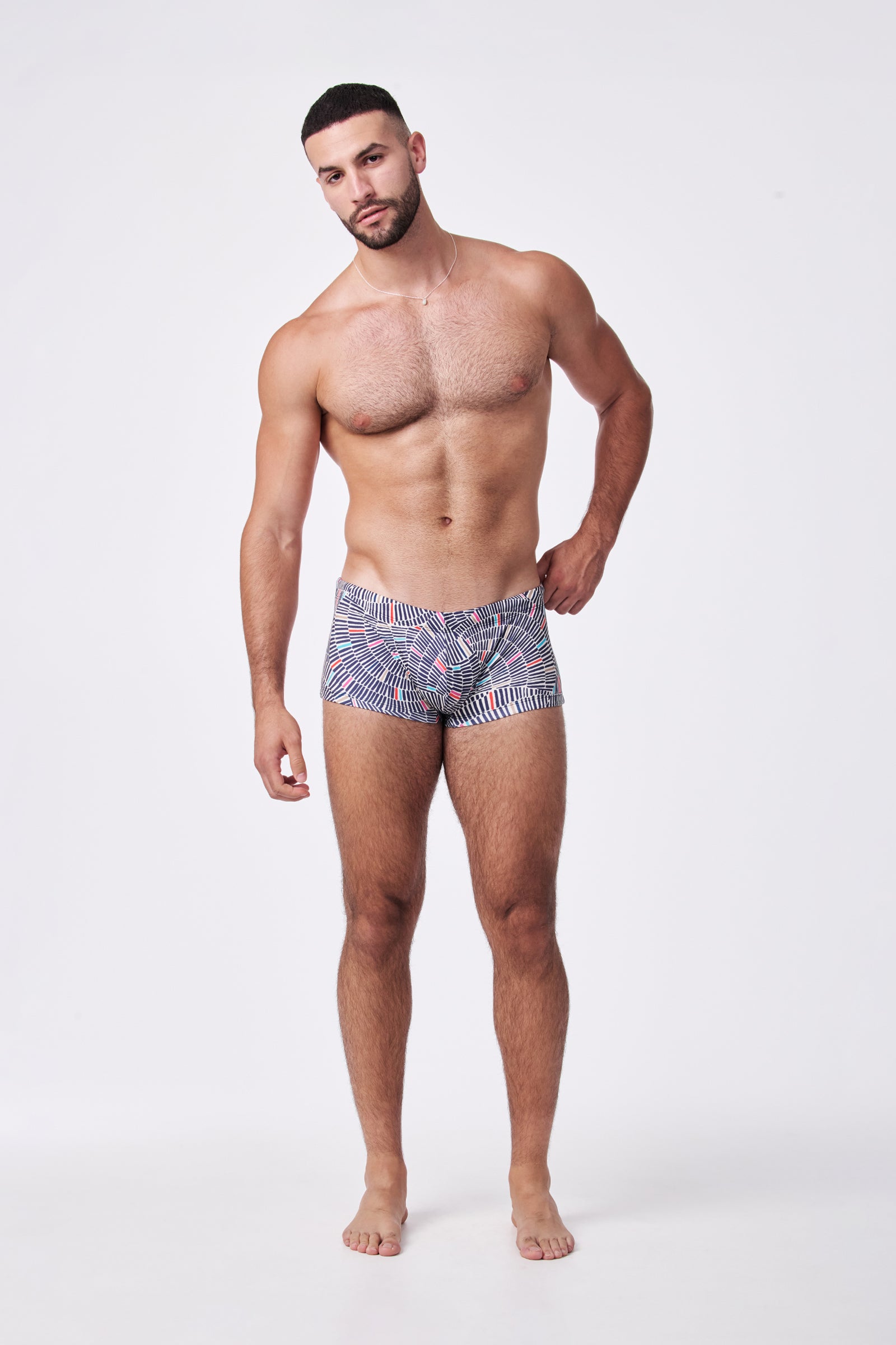 A muscular man with short dark hair and a trimmed beard poses barefoot in Sorrento Swim Trunk against a plain white background, one arm bent and the other relaxed by his side.