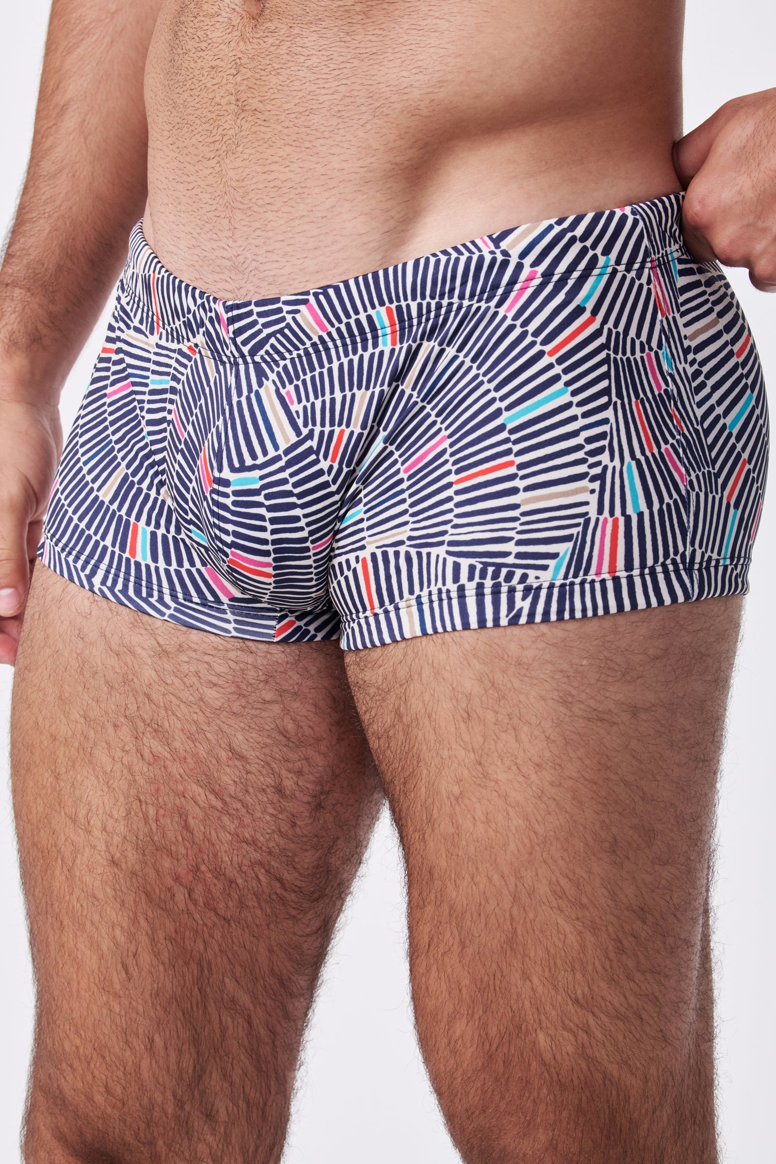 A close-up of a man wearing Sorrento Swim Trunk featuring colorful, patterned horizontal stripes and geometric shapes, standing against a plain white background.