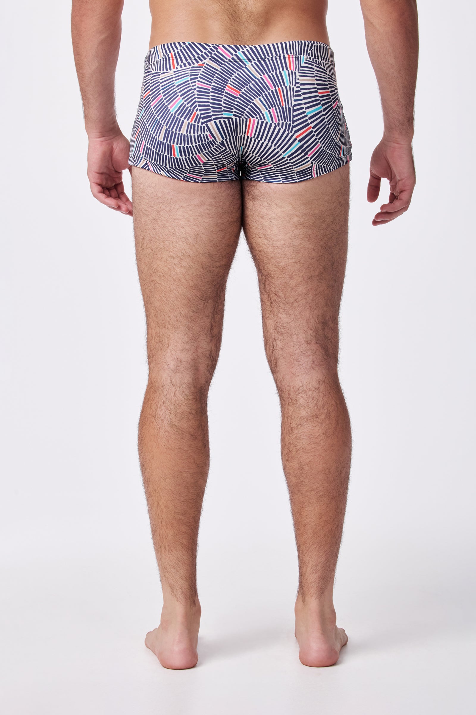 A man stands barefoot facing away, modeling the Sorrento Swim Trunk featuring a black, white, and red geometric pattern against a plain white background.