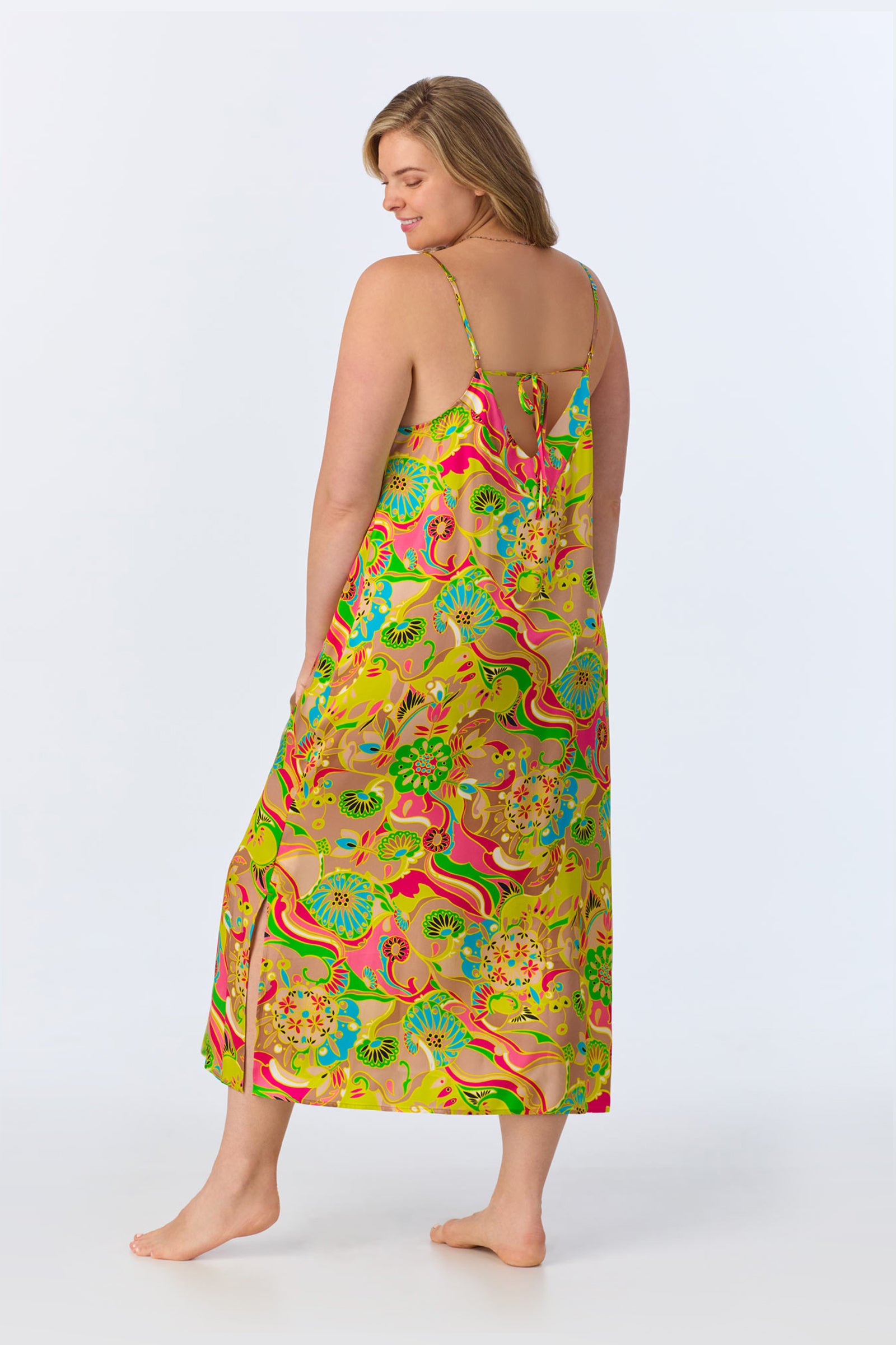 A woman stands barefoot facing away, wearing the TT Maxi Chemise Nightgown - Heritage Silk, a sleeveless, ankle-length dress with spaghetti straps and a vivid floral and swirl pattern in green, yellow, pink, blue, and orange.