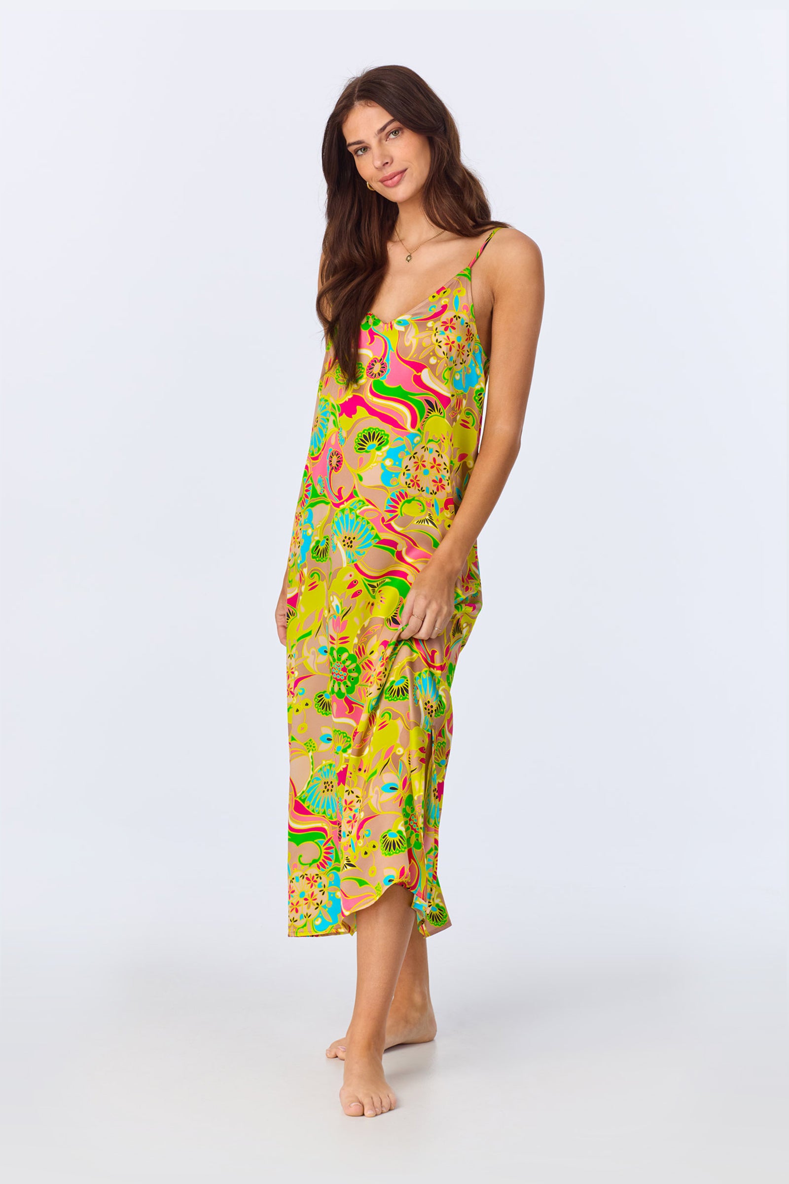 A woman with long brown hair stands barefoot against a plain white background, wearing the TT Maxi Chemise Nightgown - Heritage Silk, a colorful, sleeveless, mid-calf dress with an abstract pattern.
