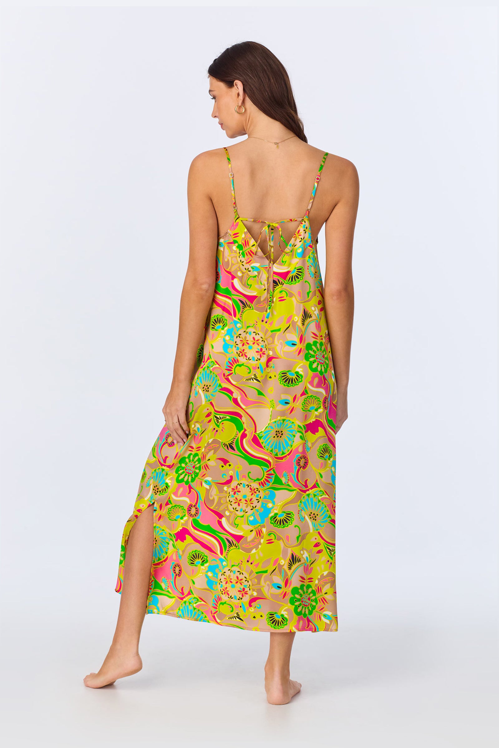 A woman stands barefoot, facing away, wearing the TT Maxi Chemise Nightgown - Heritage Silk. This colorful ankle-length dress features thin straps, a bold floral and abstract pattern in yellow, green, pink, blue, a low back, and a side slit.