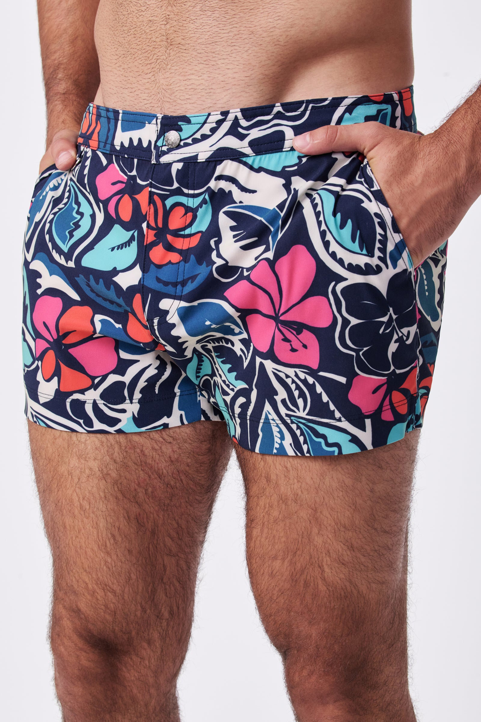 A person wearing Chico Swim Trunk - Copacabana, featuring vibrant red, pink, and blue floral prints on a dark background with hands in pockets. Photo shows only the torso and upper thighs.