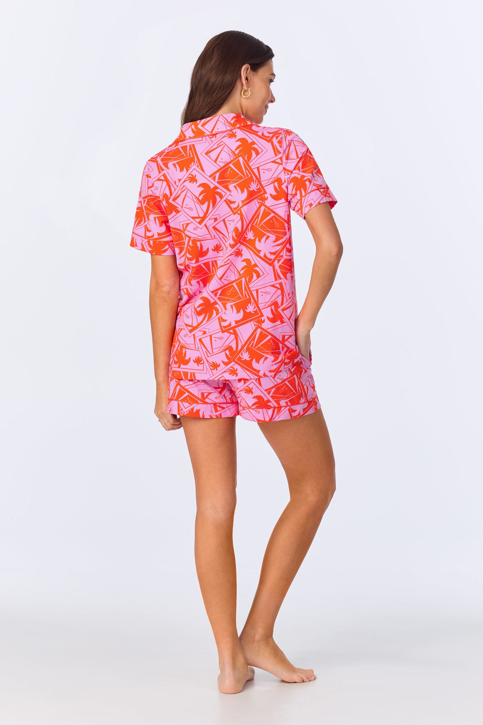 A woman with long brown hair stands barefoot, facing away, wearing the Shorty PJ Set - Tennis Stretch Jersey, featuring a matching tropical-print top with short sleeves and shorts. Her left hand is in her pocket against a plain white background.