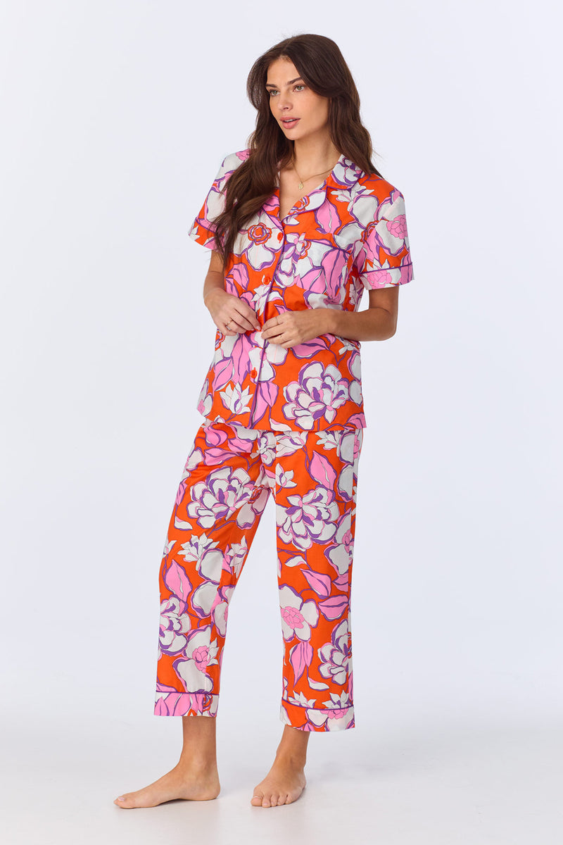A woman stands barefoot in the Crop Classic PJ Set - Gardenia Poplin, featuring vibrant floral-print poplin pajamas with a short-sleeve, button-up top and matching pants. She poses against a plain white background.