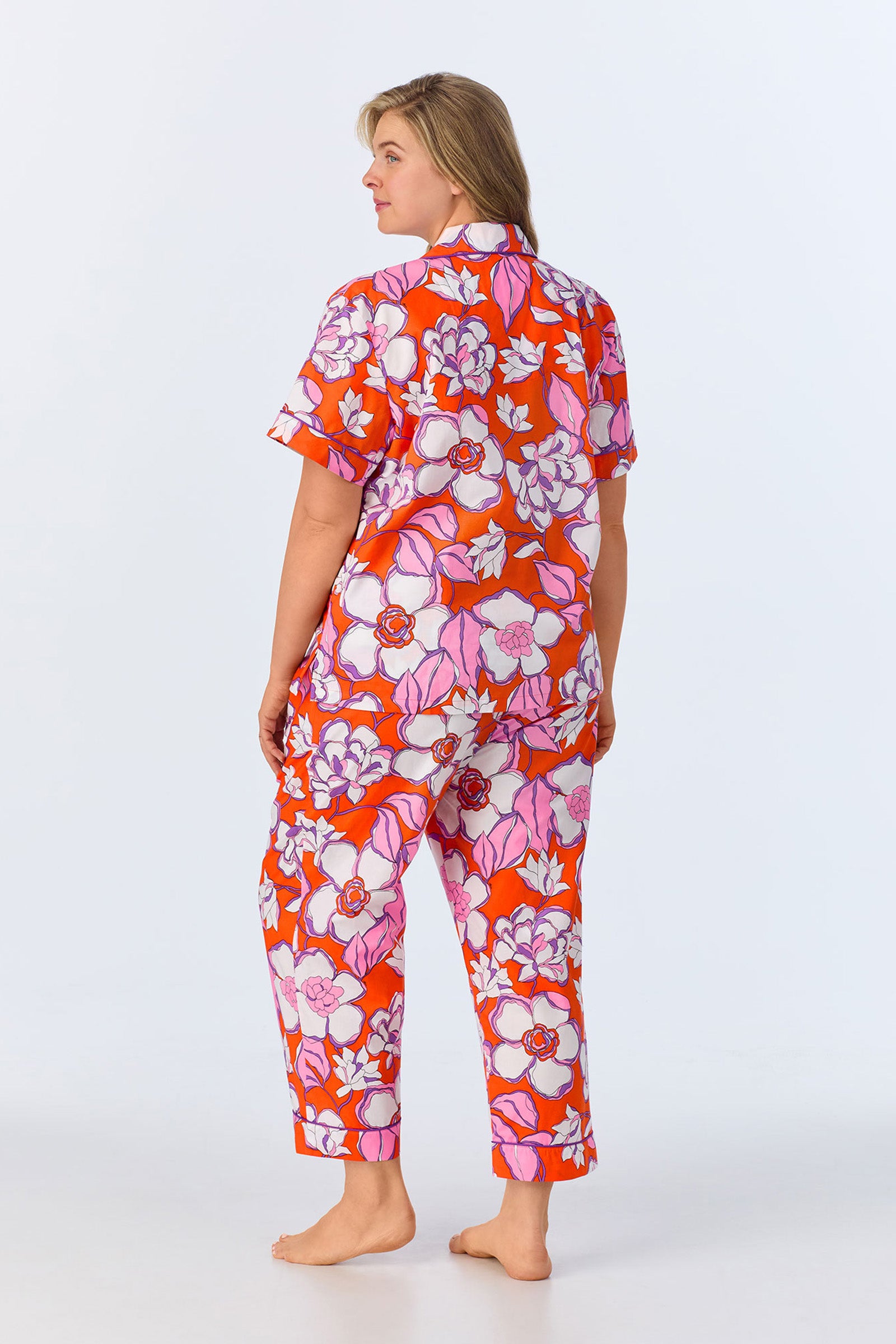 A woman stands barefoot facing away, wearing the Crop Classic PJ Set - Gardenia Poplin, which features a bold red, pink, and white floral pattern with short sleeves and long pants. The background is plain and light-colored.