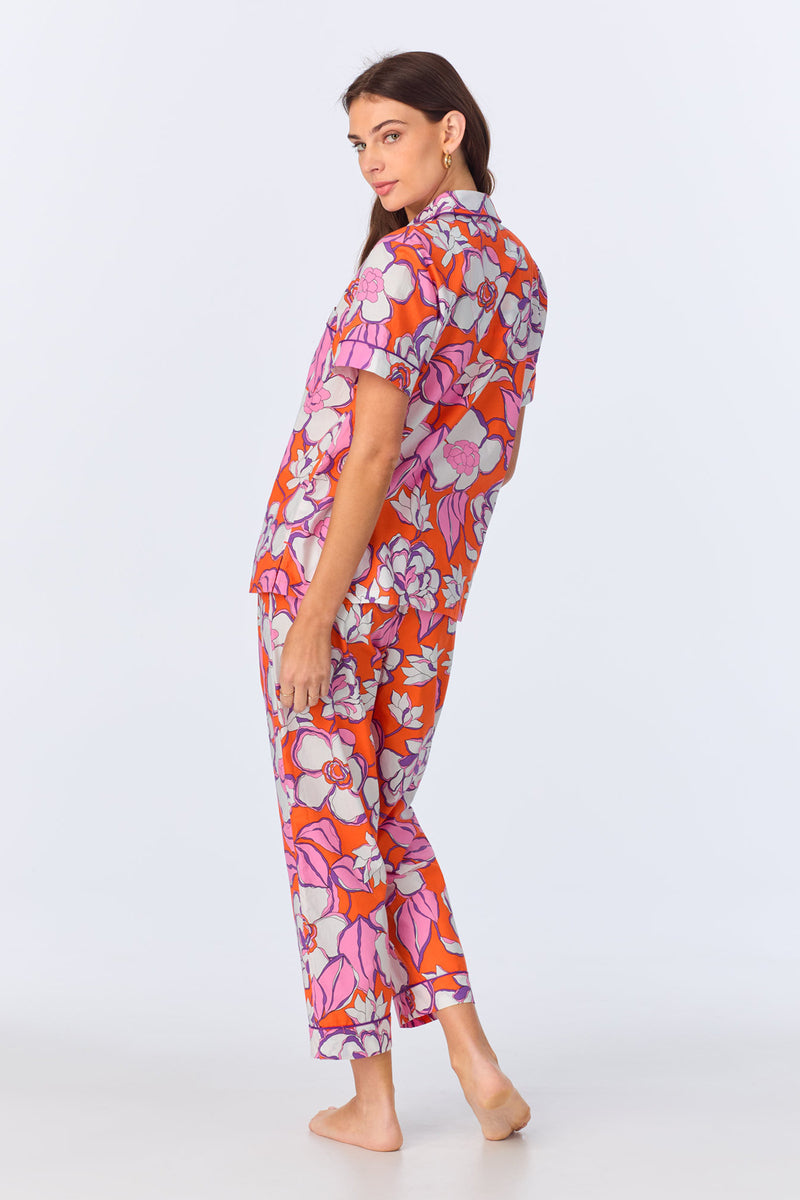 A woman stands barefoot, turned slightly to the side, wearing the Crop Classic PJ Set - Gardenia Poplin, a vibrant orange pajama set with bold pink, purple, and white florals. The set features short sleeves and cropped pants. The background is plain white.
