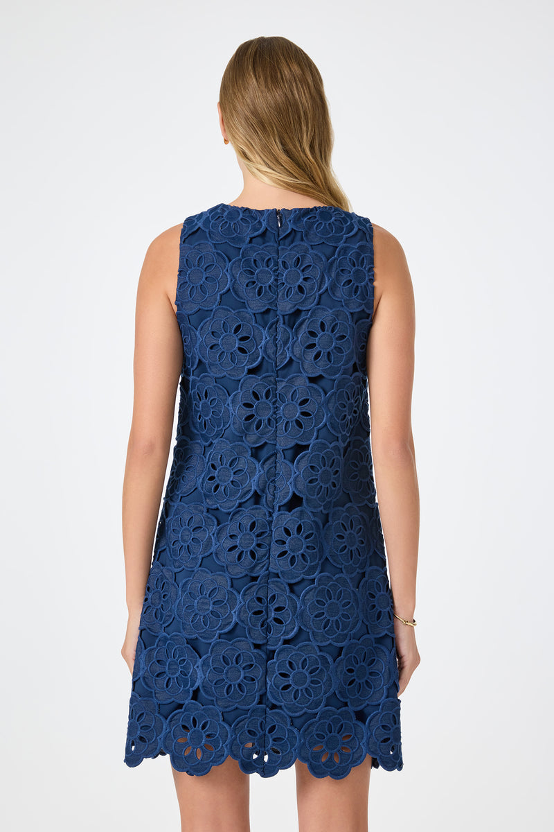 A woman with long, light brown hair faces away, modeling the Yakitori Mini Dress - Kusho Embroidery: a sleeveless, knee-length navy blue dress with large floral lace patterns and scalloped edges. The background is plain white.