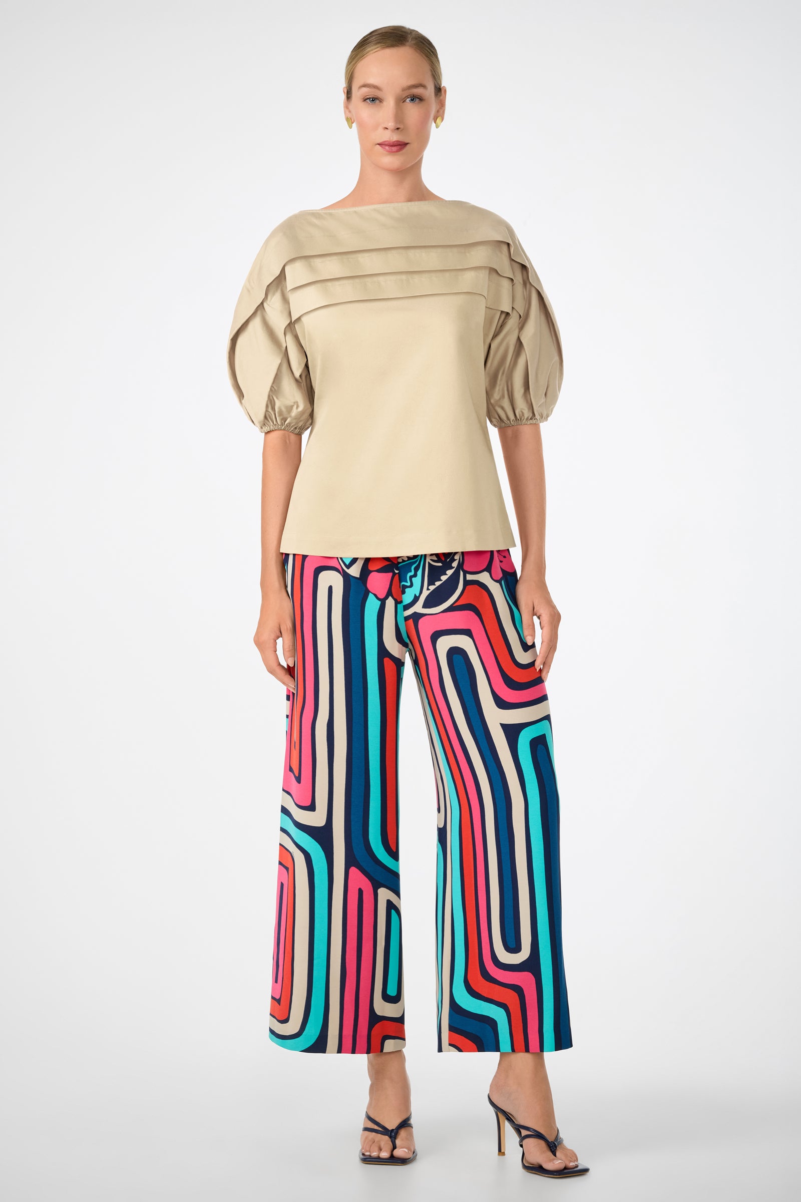 A woman wears the Grady Puff-Sleeve Top in beige with layered pleats, paired with colorful, wide-leg pants featuring a bold geometric pattern in blue, pink, red, and teal, and finishes the look with black heeled sandals.