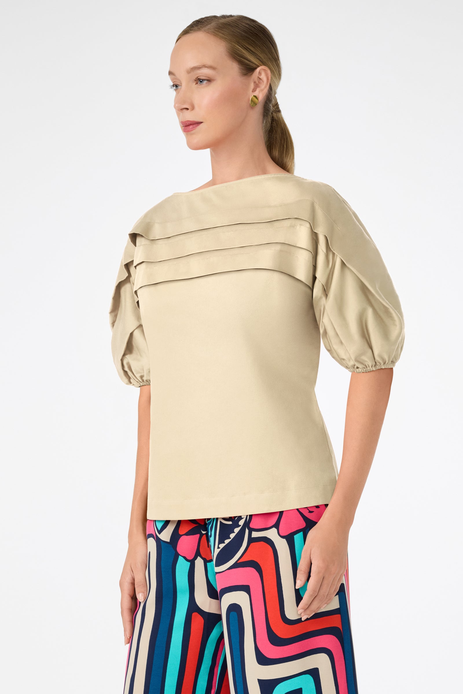 A woman wears the Grady Puff-Sleeve Top in beige, featuring pleated details and puffed half sleeves, paired with vibrant geometric-patterned pants. She stands against a plain white background, angled slightly to the side.