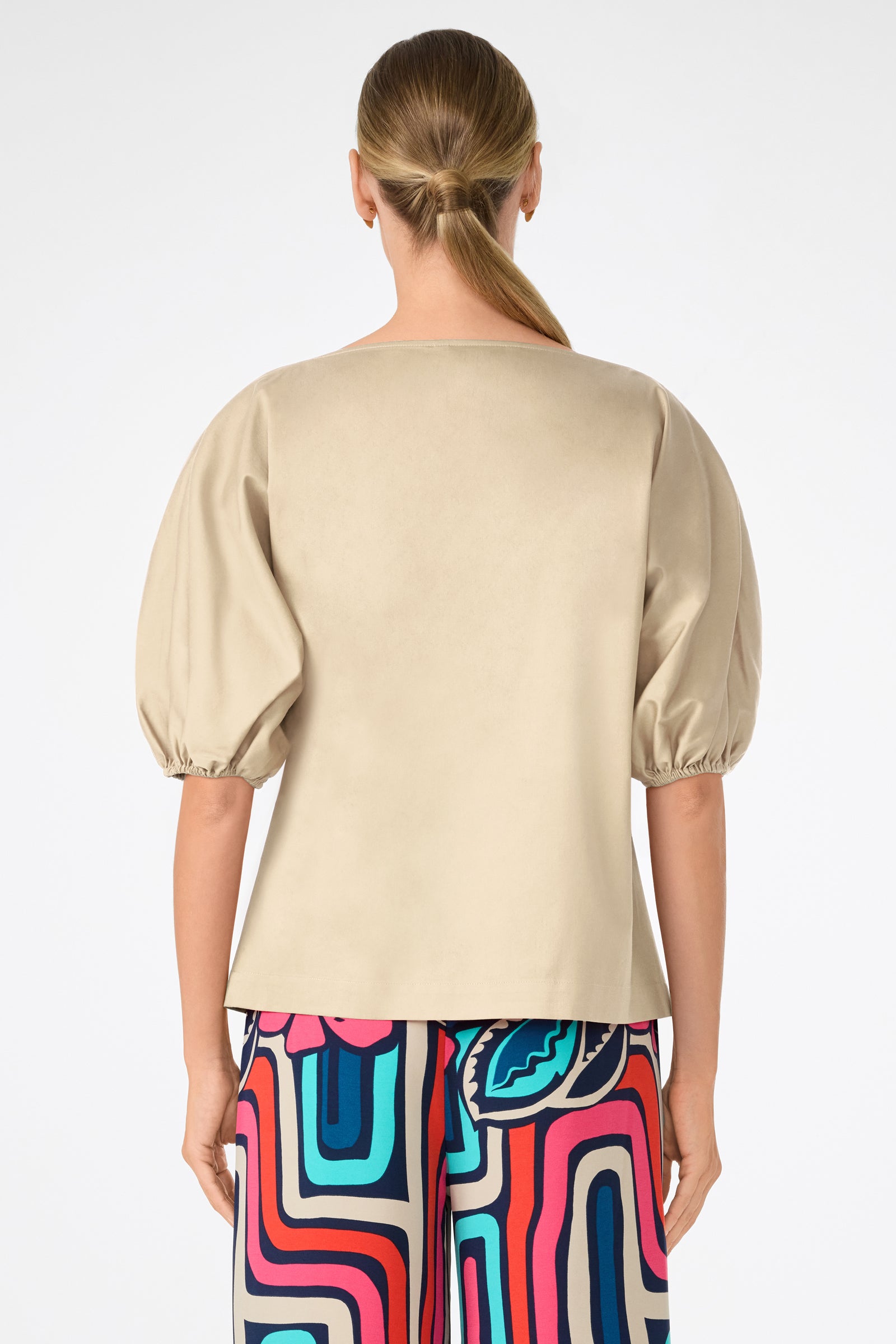 A woman with straight hair in a low ponytail is seen from behind wearing the beige Grady Puff-Sleeve Top and colorful patterned pants against a plain white background.