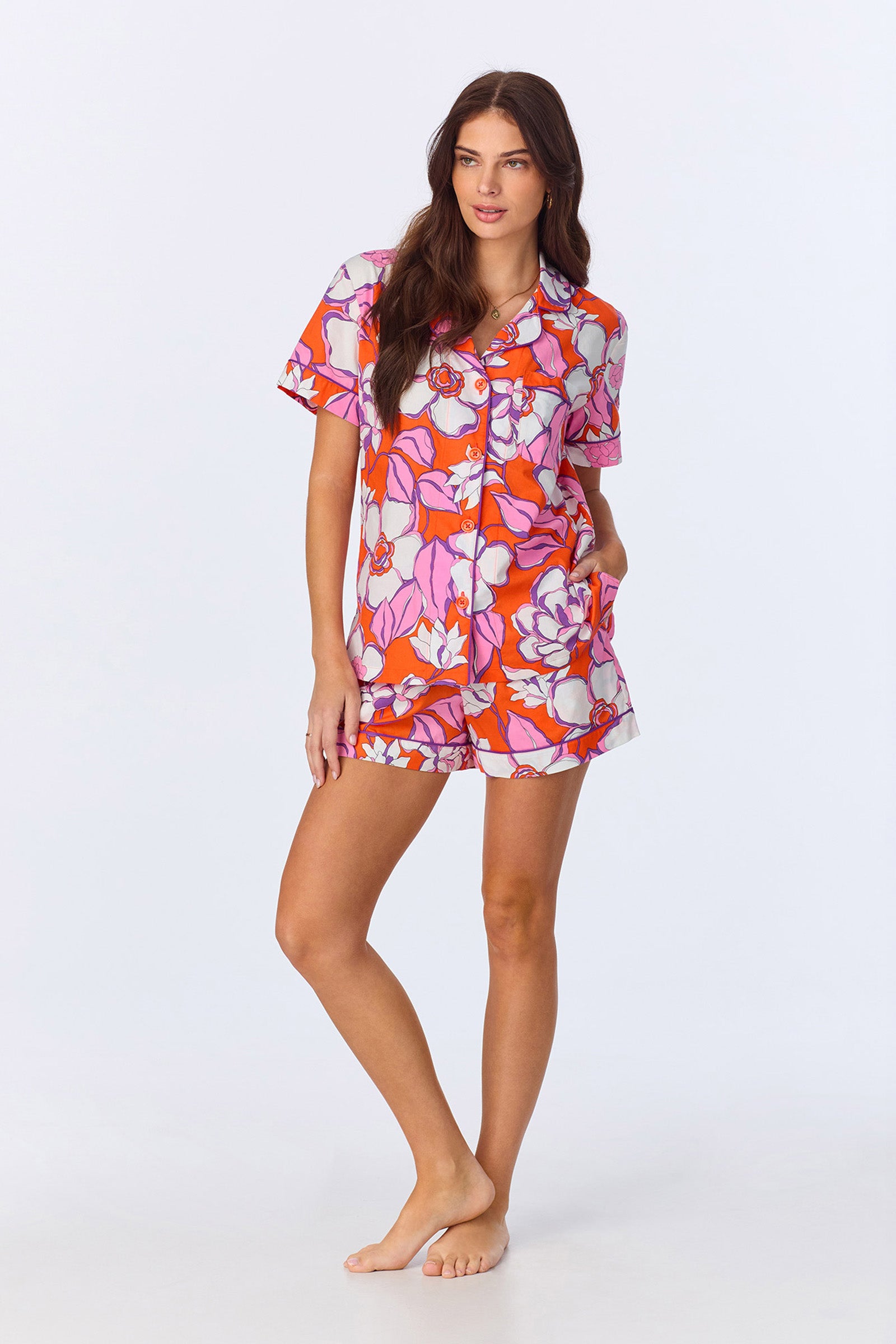 A woman stands barefoot in the Shorty PJ Set - Gardenia Poplin, featuring a multi-colored bold floral pattern. The two-piece set includes a short-sleeve button-up shirt and matching shorts.
