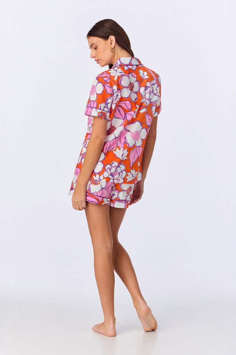A woman stands barefoot, facing slightly away, wearing the Shorty PJ Set - Gardenia Poplin featuring a bright orange, pink, and white floral print. The background is plain and light-colored.
