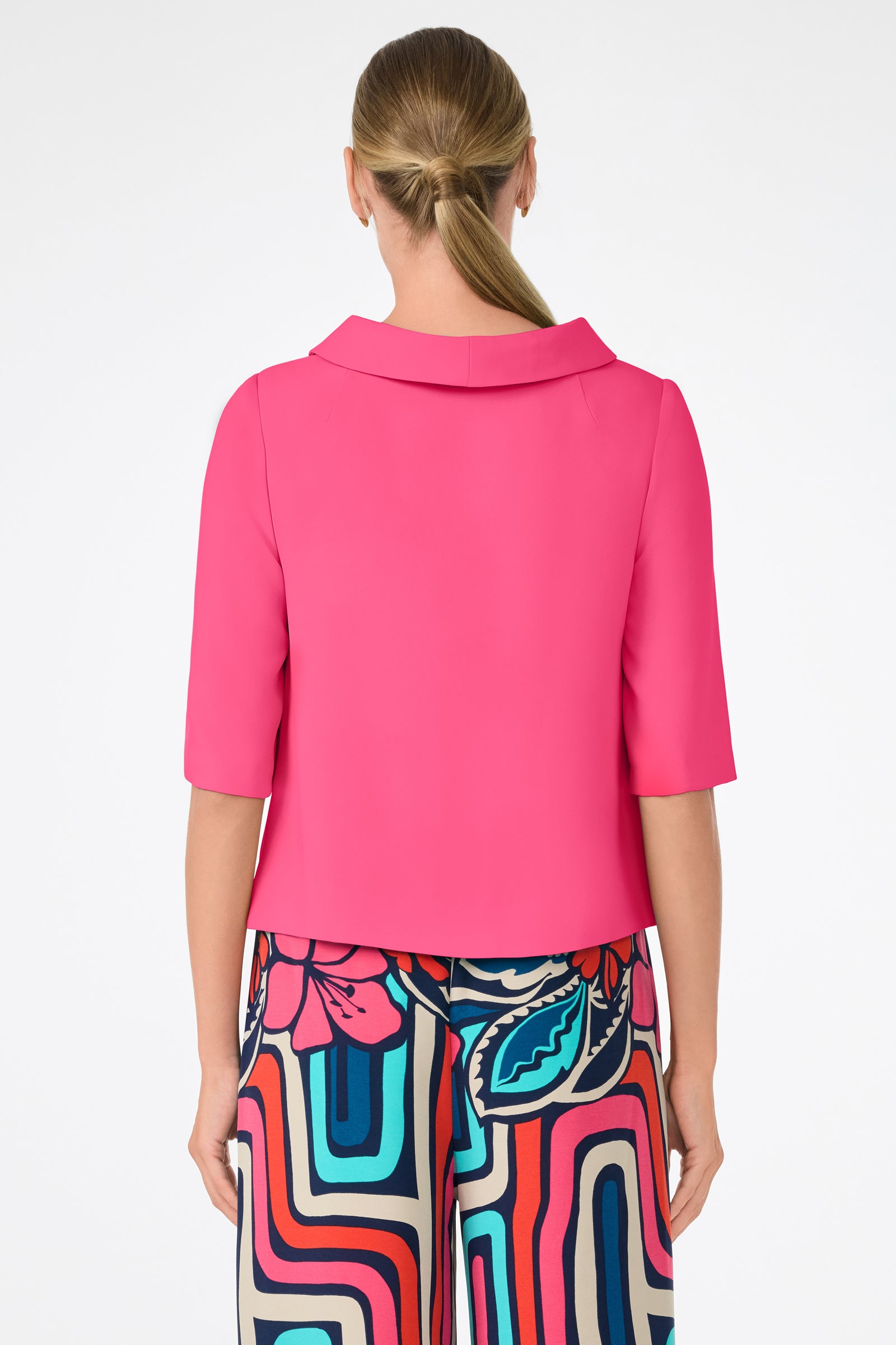 A woman with blonde hair in a low ponytail is seen from the back wearing the Kailee Top, a bright pink short-sleeve top, paired with colorful high-waisted pants featuring abstract and floral patterns against a white background.