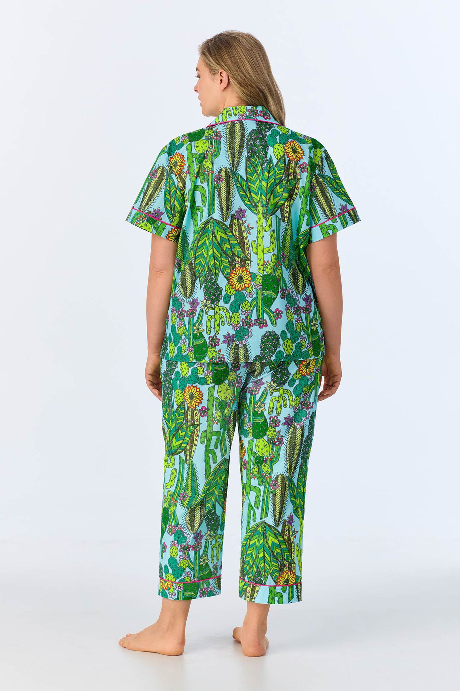 A woman with long blonde hair stands barefoot, facing away, wearing the Crop Classic PJ in Cactus Poplin—a matching pajama set featuring a bright green jungle leaf and animal print design on a light background.