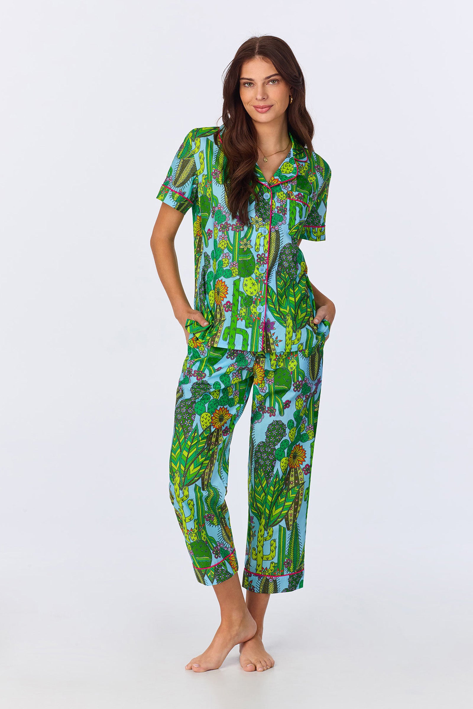 A woman with long brown hair stands barefoot against a plain background, wearing the Crop Classic PJ - Cactus Poplin set in Multi, featuring a vibrant botanical and animal print. Her hands are in her pockets.