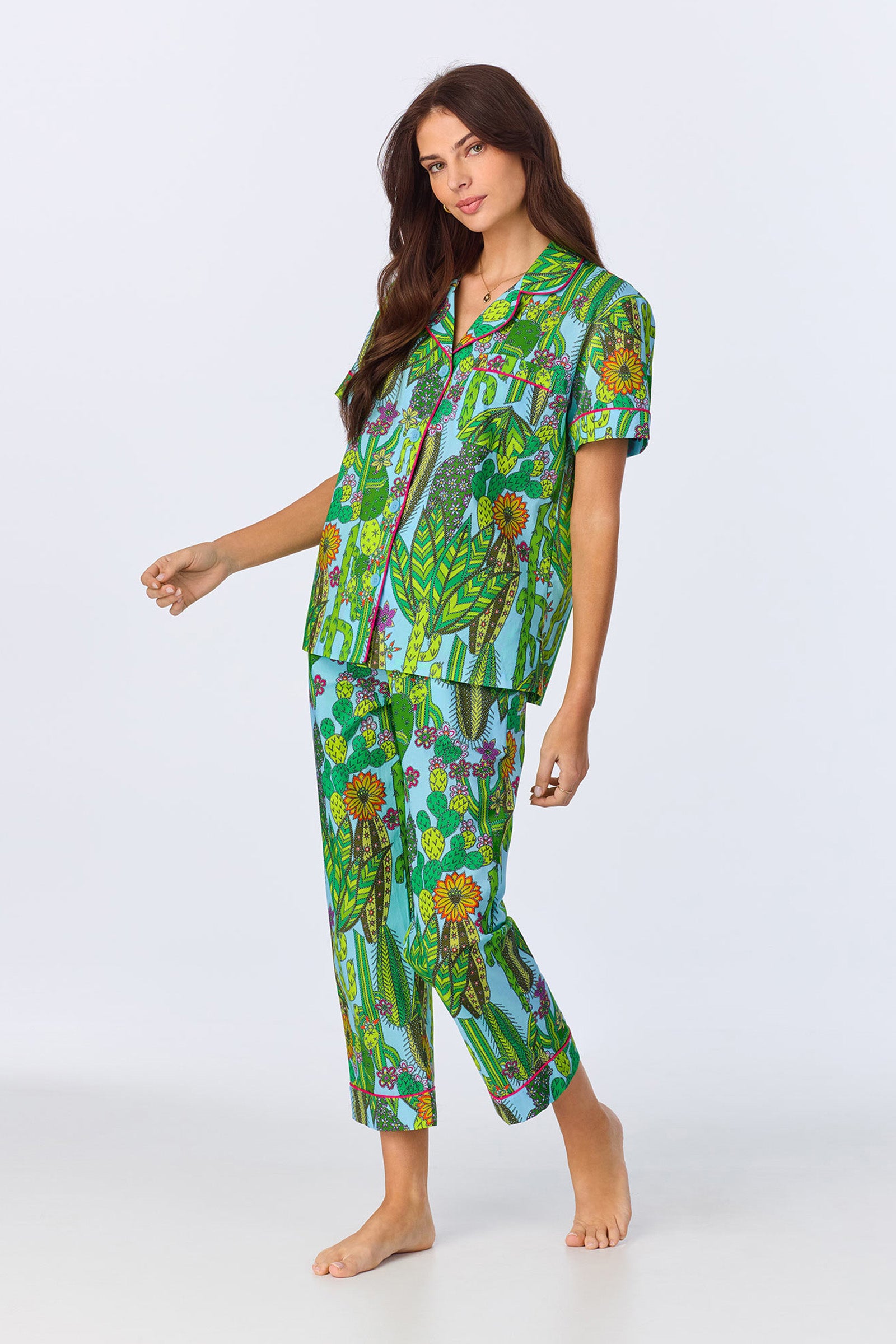 A woman with long brown hair smiles barefoot in the Crop Classic PJ - Cactus Poplin, featuring a colorful floral print, against a plain light background.