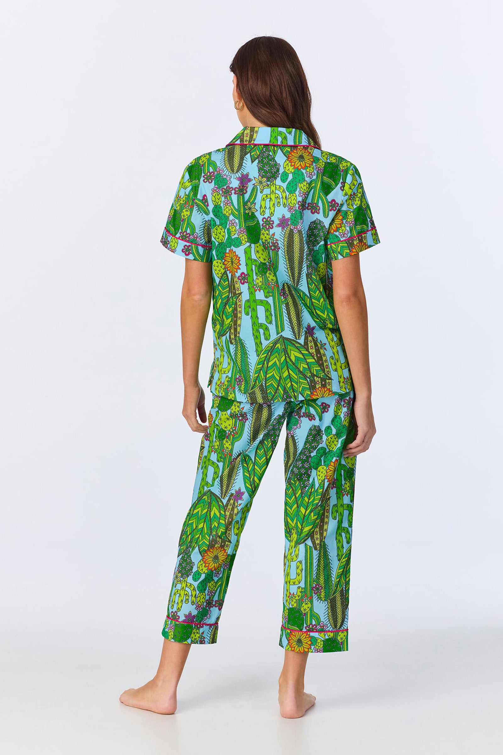A person with long brown hair stands barefoot facing away, wearing the Crop Classic PJ - Cactus Poplin, a matching pajama set with vibrant green, blue, and yellow botanical print, featuring short sleeves and cropped pants.