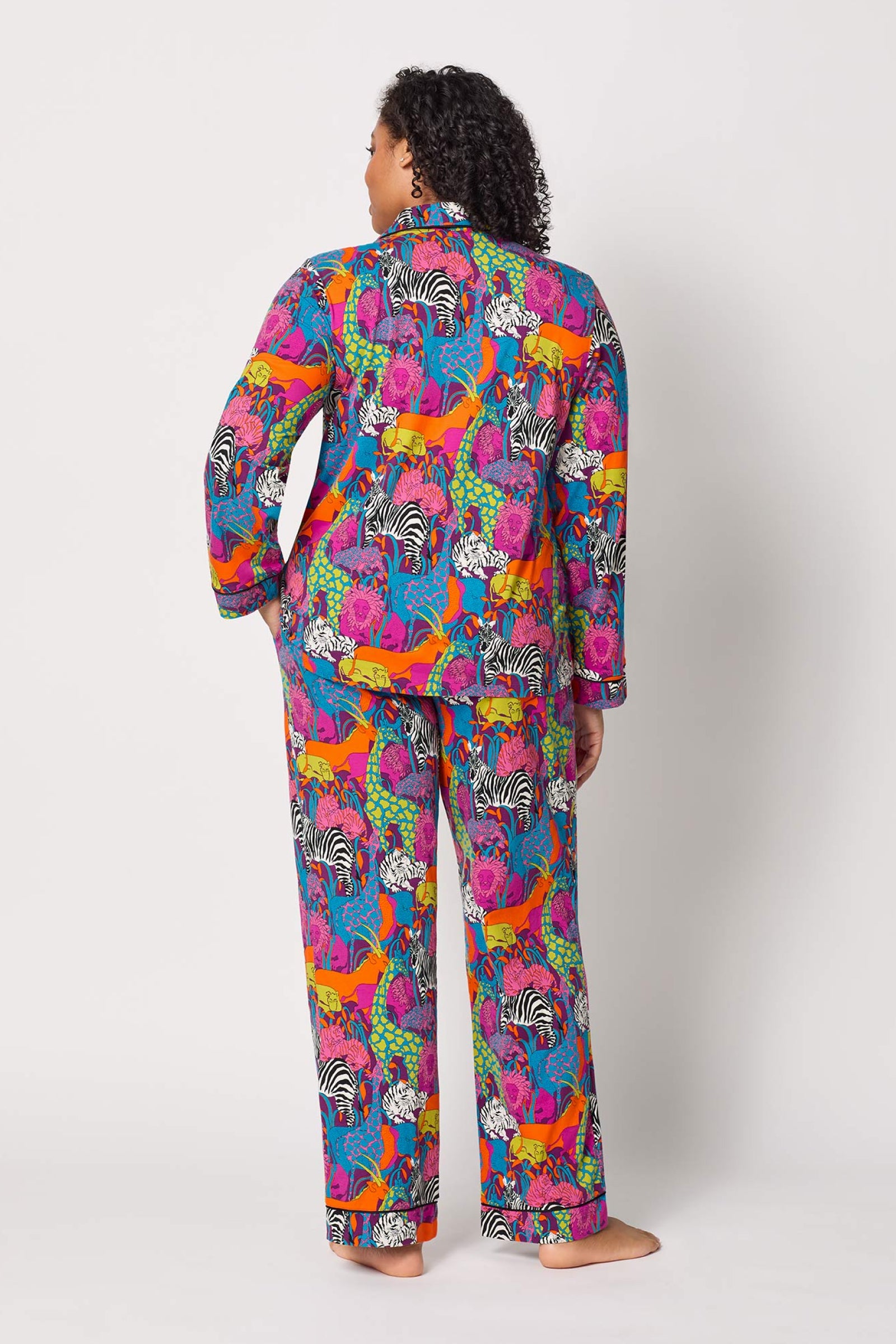 A person stands barefoot, facing away, wearing the Classic PJ Set - Jungle, featuring a vibrant animal and floral print in blue, pink, orange, green, and yellow on a white background.