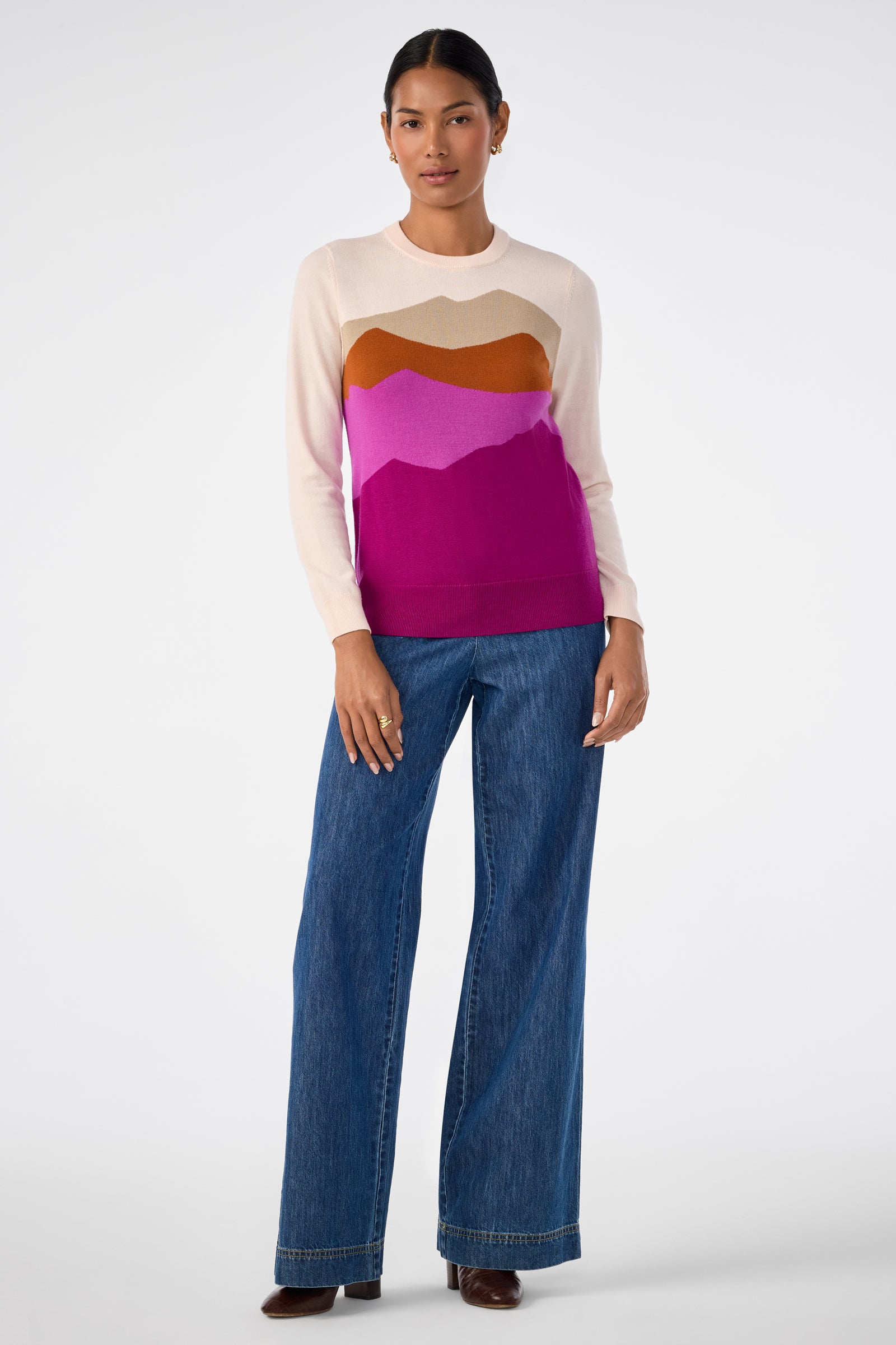 A woman faces forward against a white background, wearing the Jacinto Crewneck—a long-sleeve sweater with layered cream, brown, and pink—and wide-leg blue jeans with brown shoes. One hand rests by her side.