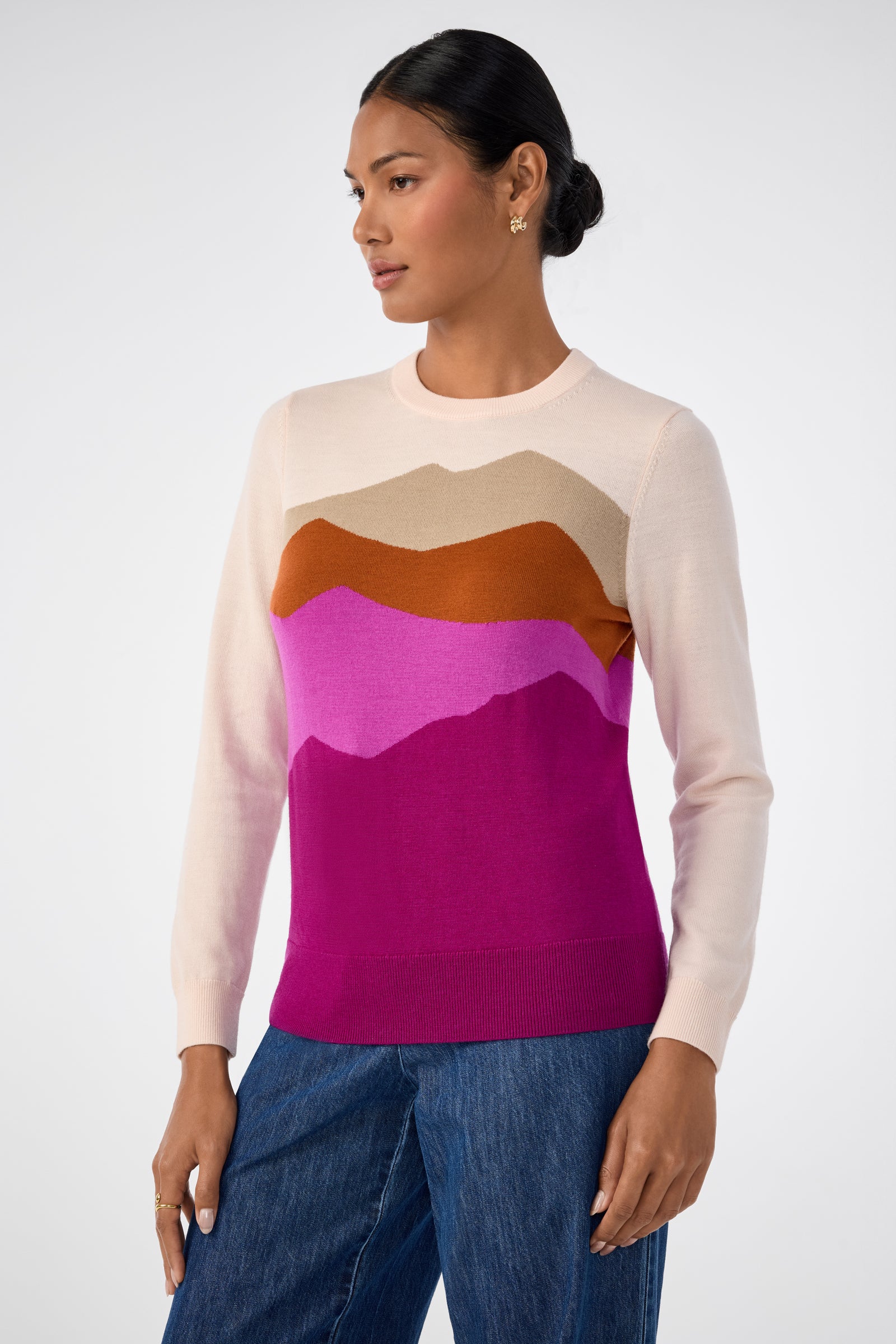 A woman with dark hair in a low bun models the Jacinto Crewneck, a pastel sweater featuring a colorful mountain pattern in tan, orange, pink, and purple. She pairs it with blue jeans and looks to the side against a plain background.
