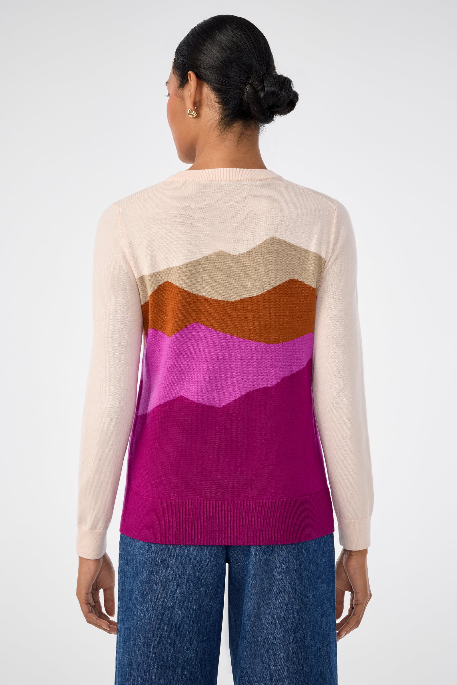 A woman with dark hair in a low bun, seen from behind, wears the Jacinto Crewneck—a light sweater featuring a mountain-inspired pattern in brown, tan, pink, and magenta—paired with wide-leg blue jeans.