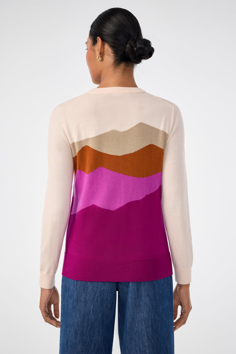 A woman with dark hair in a low bun, seen from behind, wears the Jacinto Crewneck—a light sweater featuring a mountain-inspired pattern in brown, tan, pink, and magenta—paired with wide-leg blue jeans.