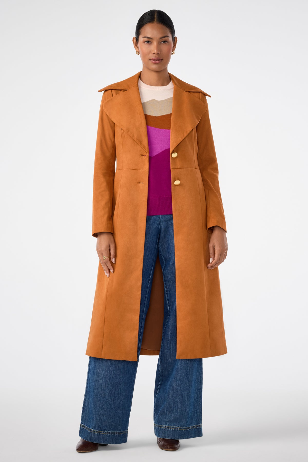 A person stands facing forward in a Jacinto Crewneck, an orange-brown long coat, wide-leg blue jeans, and brown shoes against a plain white background.