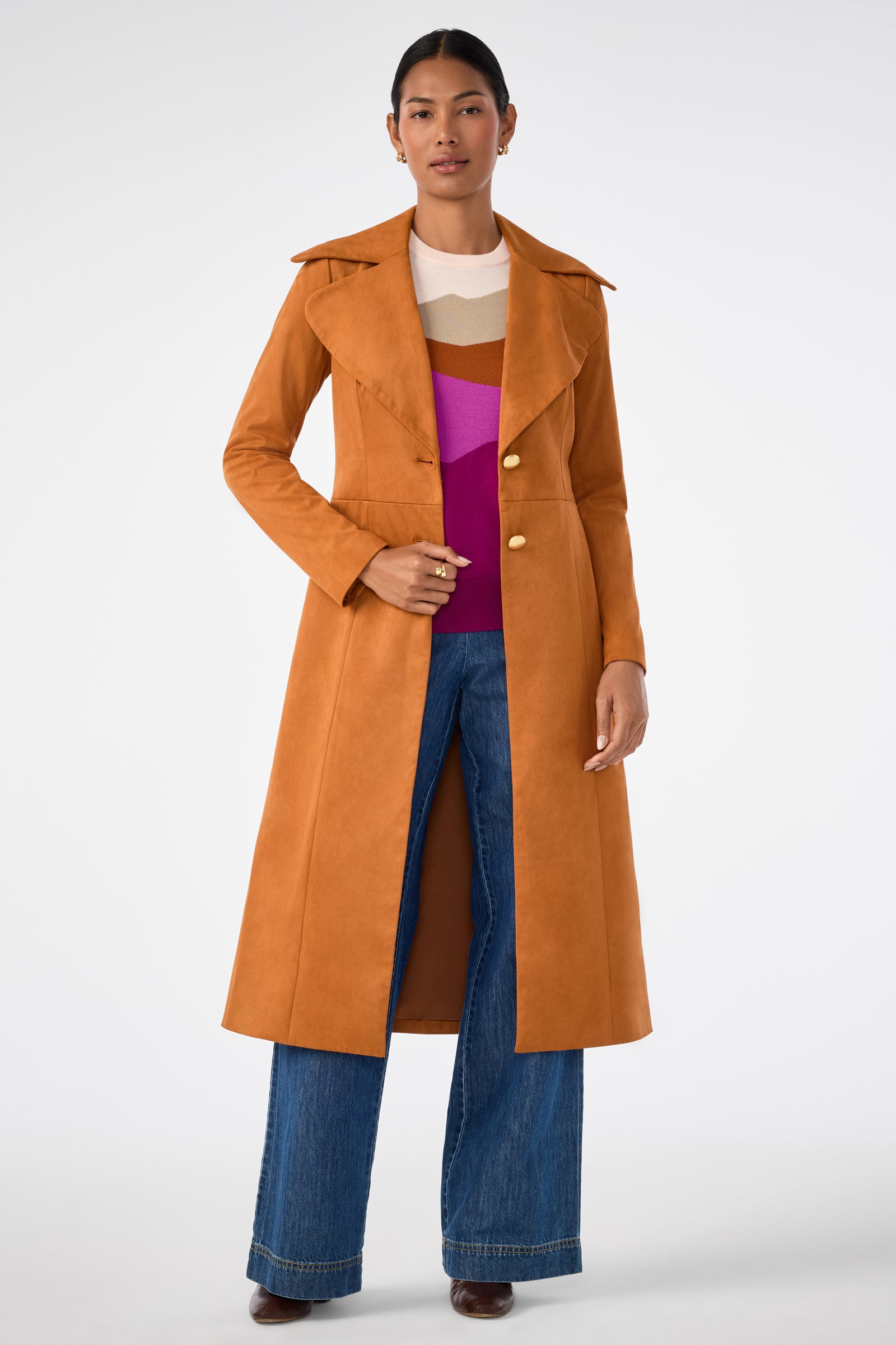 A woman stands facing forward, wearing the Starflower Coat—a long, camel trench coat with gold buttons—over a color-blocked top and wide-leg blue jeans. Her dark hair is pulled back and she pairs the look with brown heeled shoes.