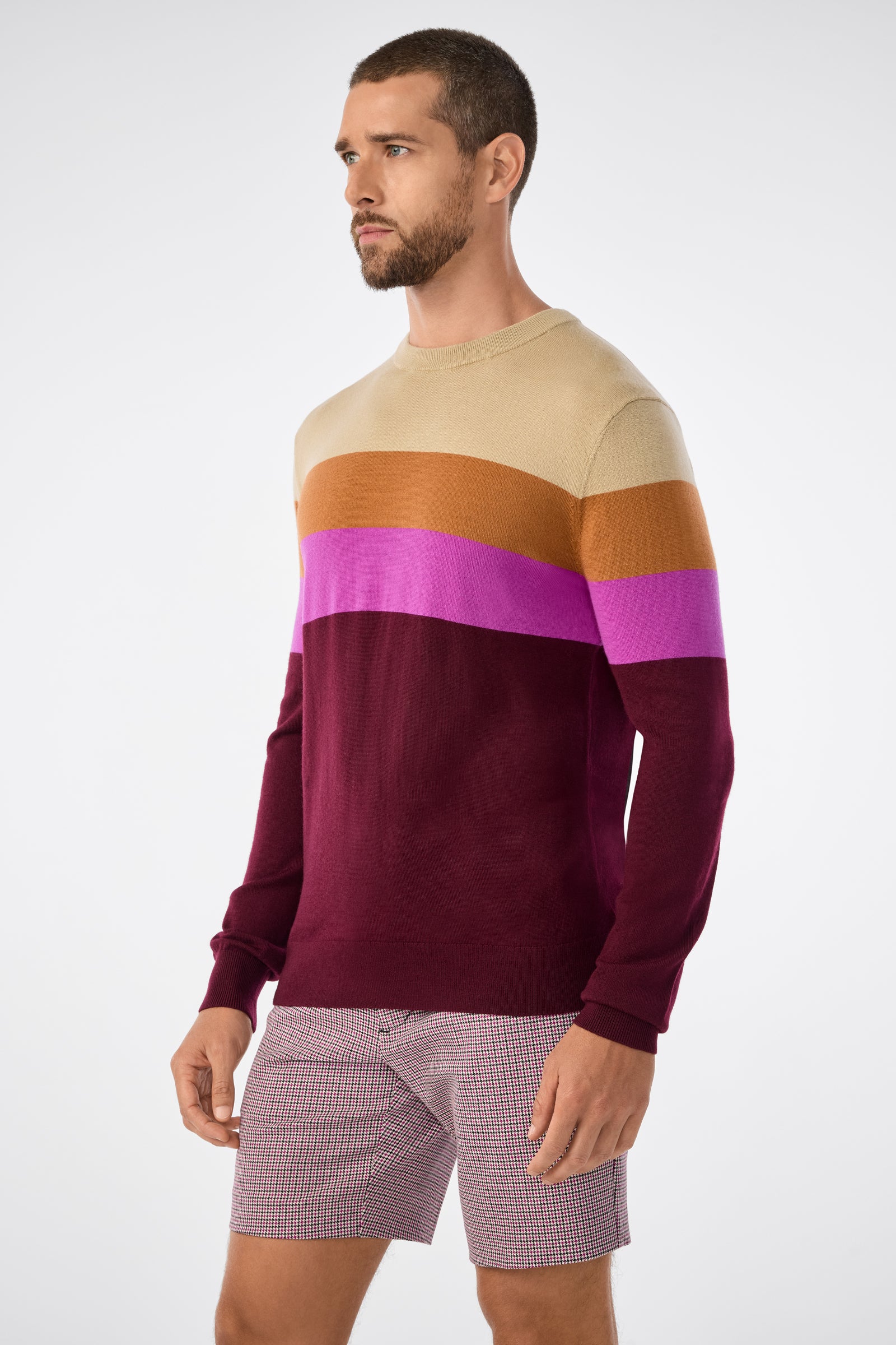 A man with short hair and a beard wears the Romero Sweater, featuring beige, orange, purple, and burgundy color-block stripes, paired with red-and-white checkered shorts, standing against a light gray background.