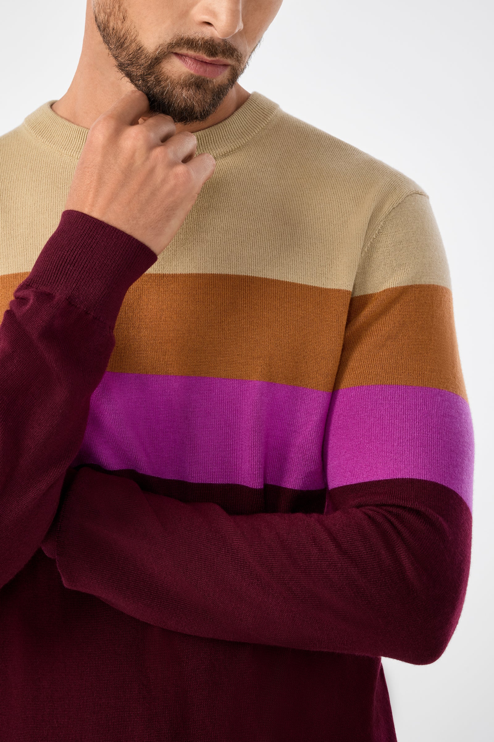 A man with a short beard wears the Romero Sweater, featuring color-block stripes in beige, brown, purple, and burgundy. He rests his chin on his hand while crossing his other arm over his chest.
