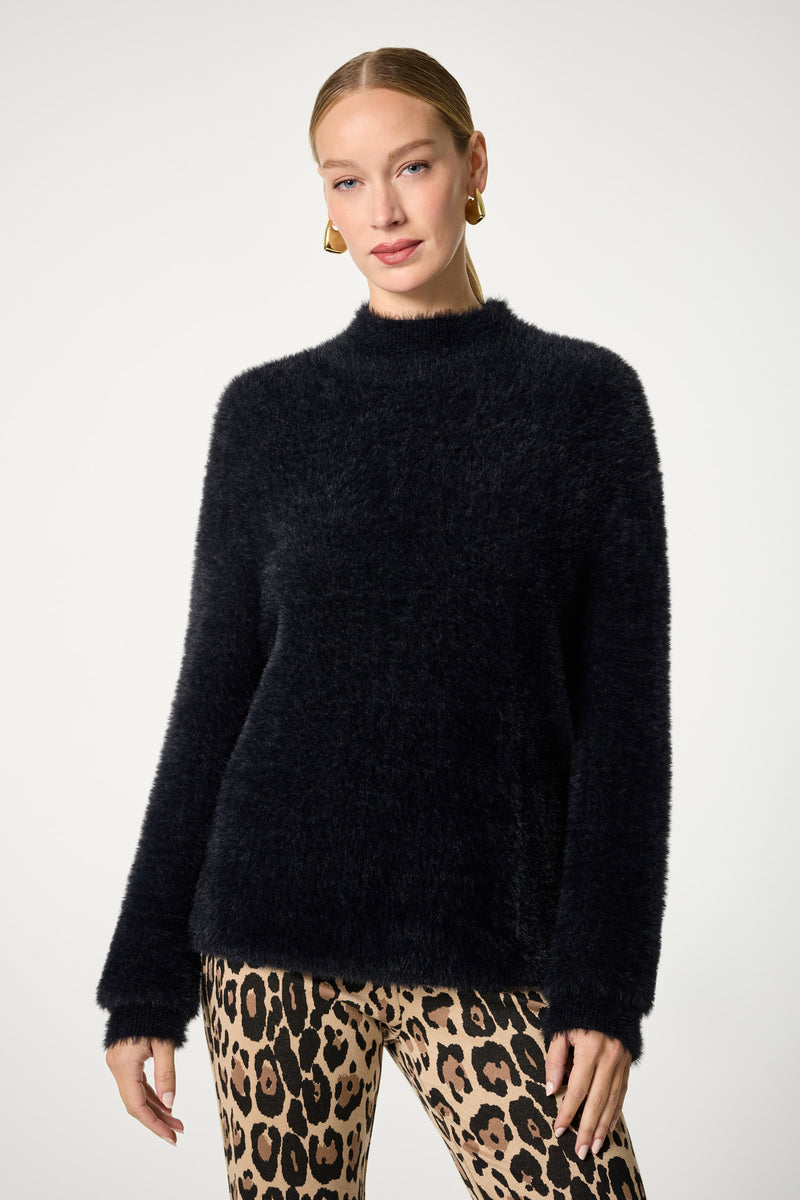 A woman stands against a plain background, wearing the Bazaar Mock Neck Sweater in black with long sleeves and fuzzy texture, paired with leopard print pants. She accessorizes with gold hoop earrings and has her hair pulled back.