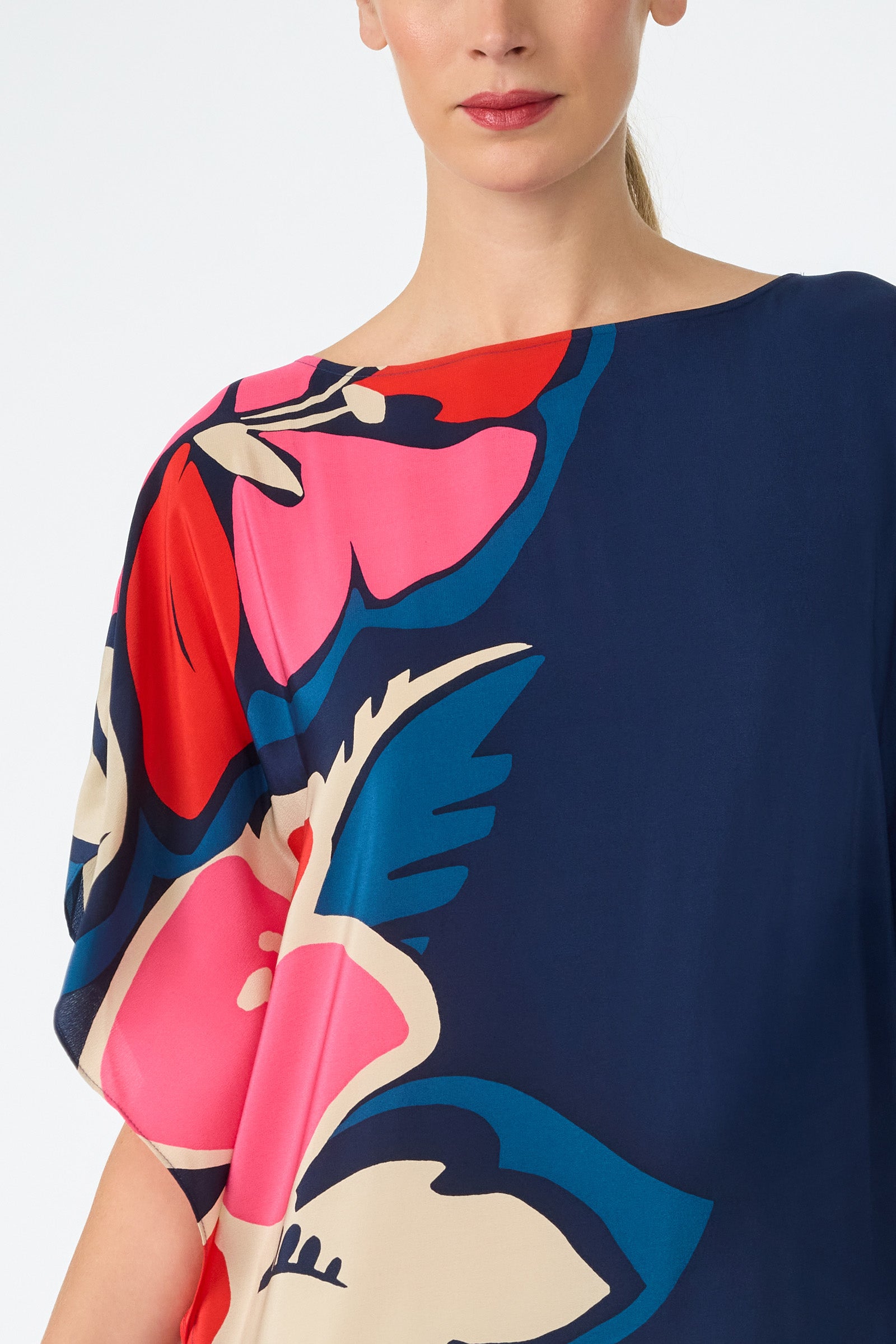 A woman wears the Jalani Maxi Caftan Dress, featuring a navy blue base with large, bold floral prints in pink, red, cream, and blue. She's shown from the shoulders up with part of her face visible and her hair tied back.