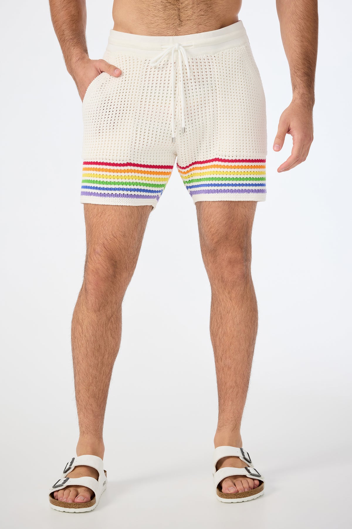 Wearing the August Open-Knit Short - Pride in Multi, paired with white slide sandals, a person stands against a plain white background, showing only their lower torso and legs. Rainbow stripes accent the cream knit shorts near the hem.