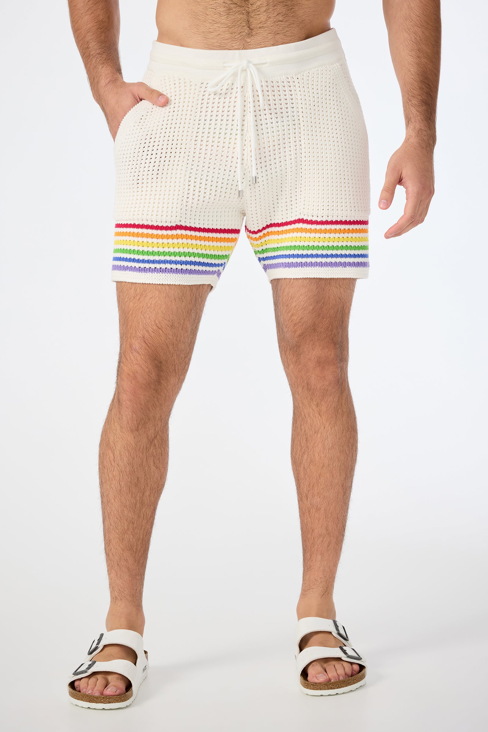 Wearing the August Open-Knit Short - Pride in Multi, paired with white slide sandals, a person stands against a plain white background, showing only their lower torso and legs. Rainbow stripes accent the cream knit shorts near the hem.